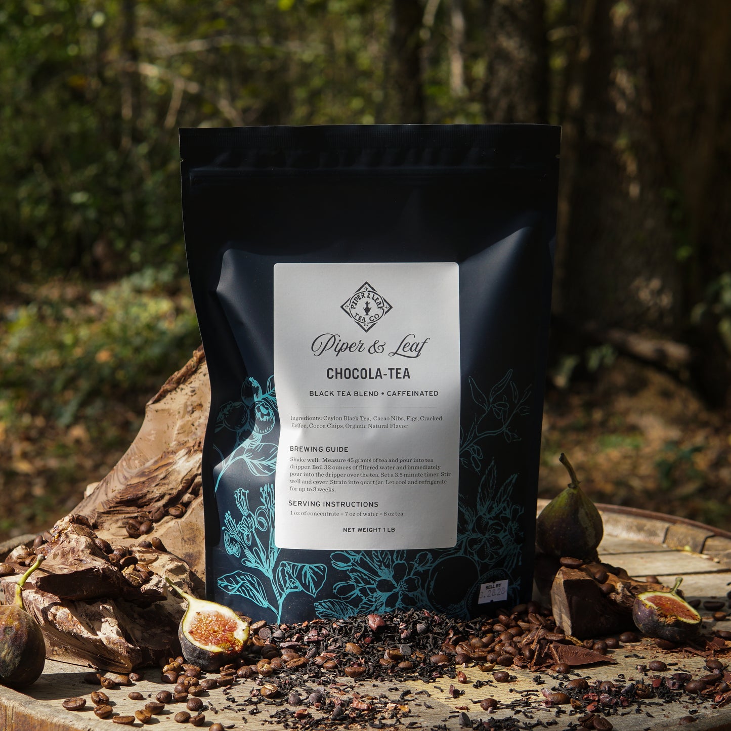 A Chocola-Tea Pound Bag (190 servings) from Piper & Leaf Tea Co. rests on a wooden surface outdoors, surrounded by figs, cacao nibs, and cocoa pods.