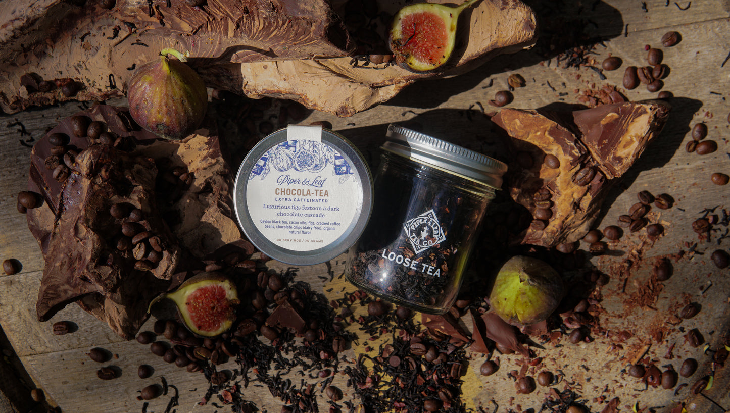 A Chocola-Tea Glass Jar of Loose Leaf Tea by Piper & Leaf Tea Co., chocolate pieces, figs, and scattered coffee beans are arranged on a wooden surface.