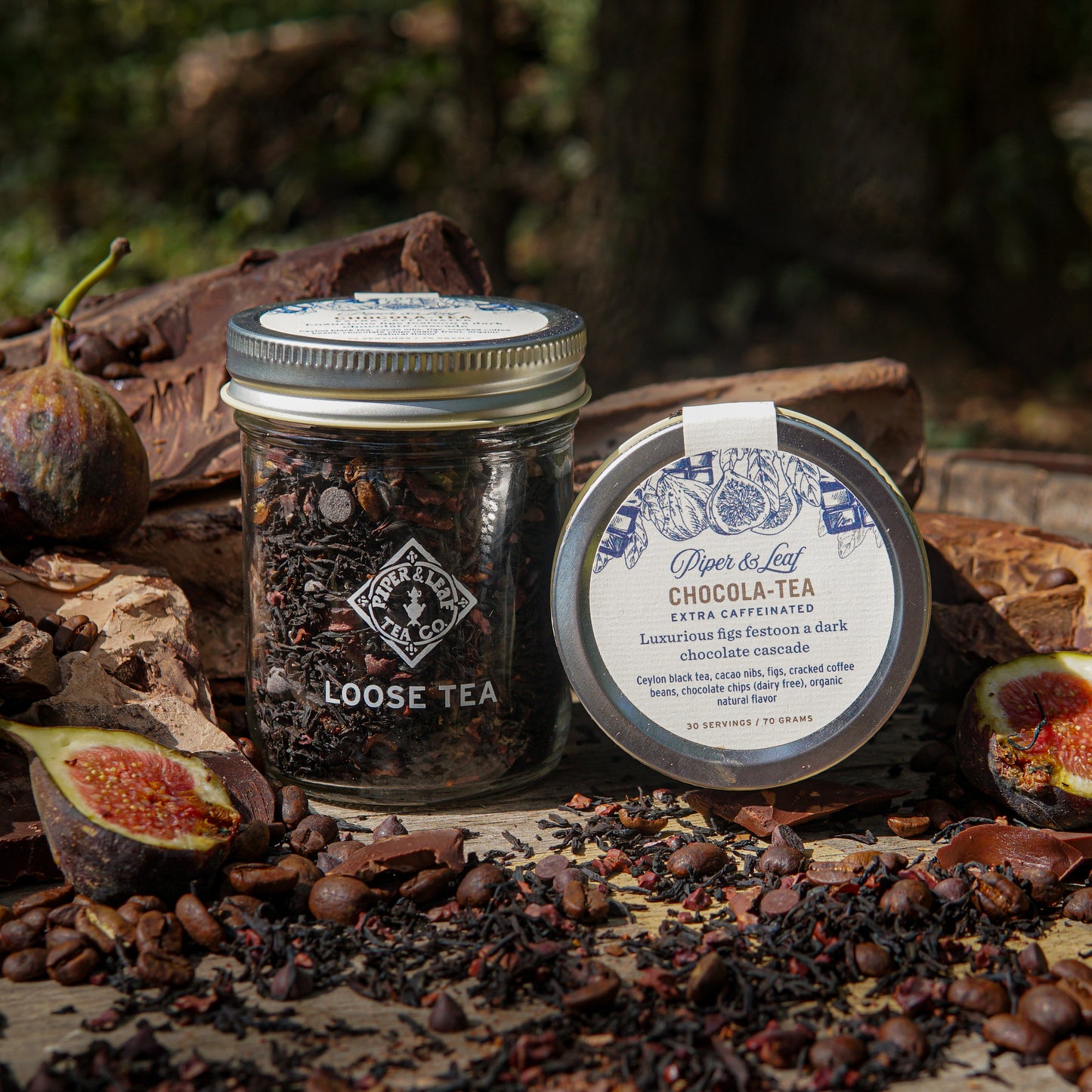 A Chocola-Tea Glass Jar of Loose Leaf Tea by Piper & Leaf Tea Co. sits amid figs, cocoa beans, and chocolate, showcasing this delicious chocolate fig tea blend with 30 servings of caffeinated tea.