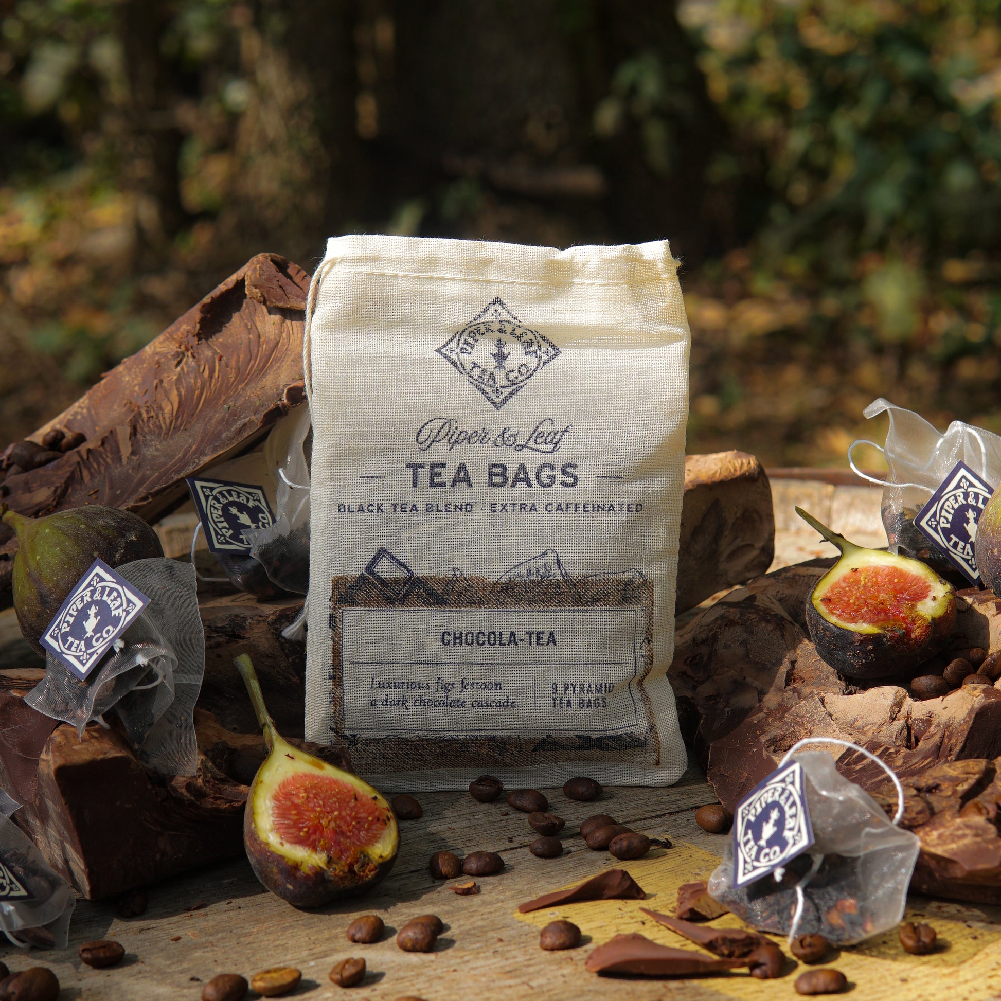 A bag of Piper & Leaf Tea Co. Chocola-Tea 9ct Tea Bags in Muslin rests on a wooden surface, surrounded by figs, dark chocolate pieces, coffee beans, and pyramid tea sachets.