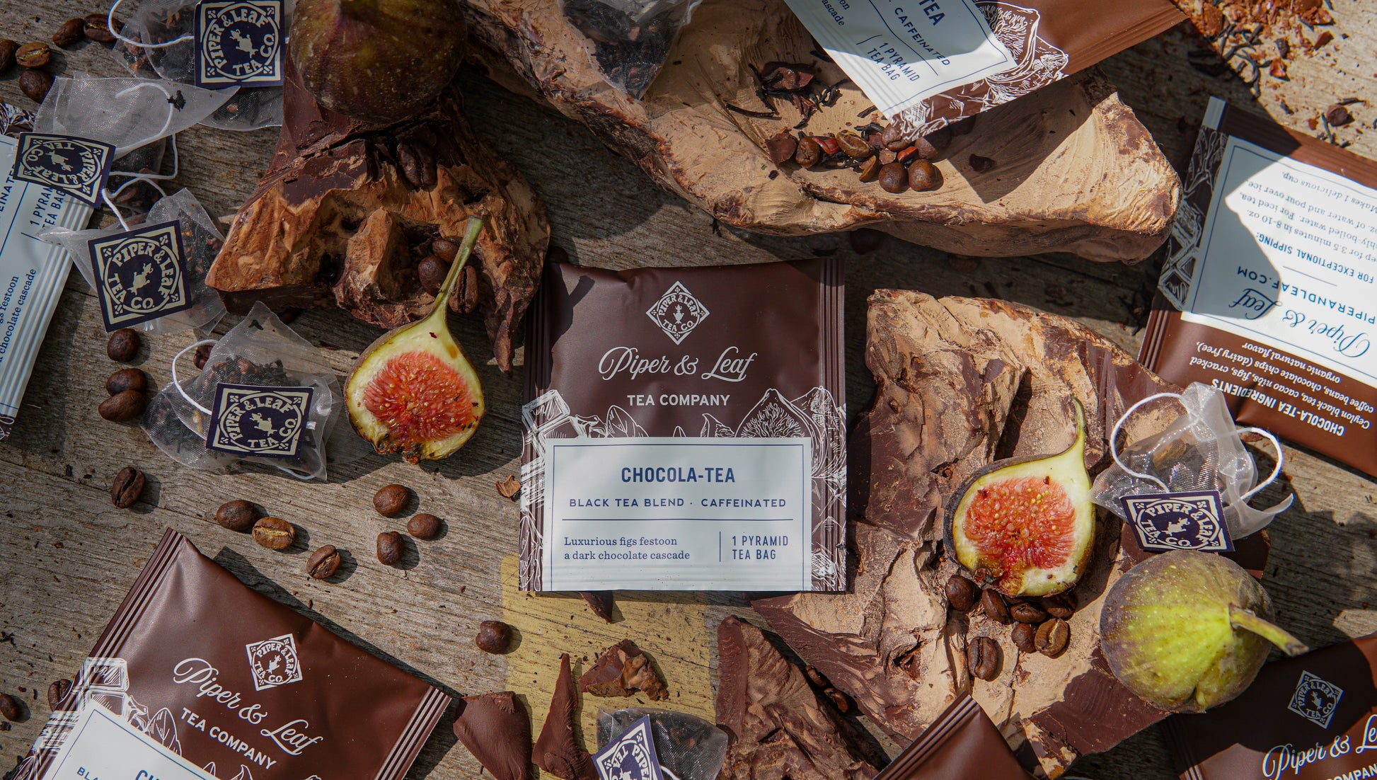 Piper & Leaf Tea Co. Chocola-Tea Case of Individually Wrapped Tea Bags (50ct) sits on a rustic wooden surface, surrounded by figs, cocoa pieces, coffee beans, and tea bags.