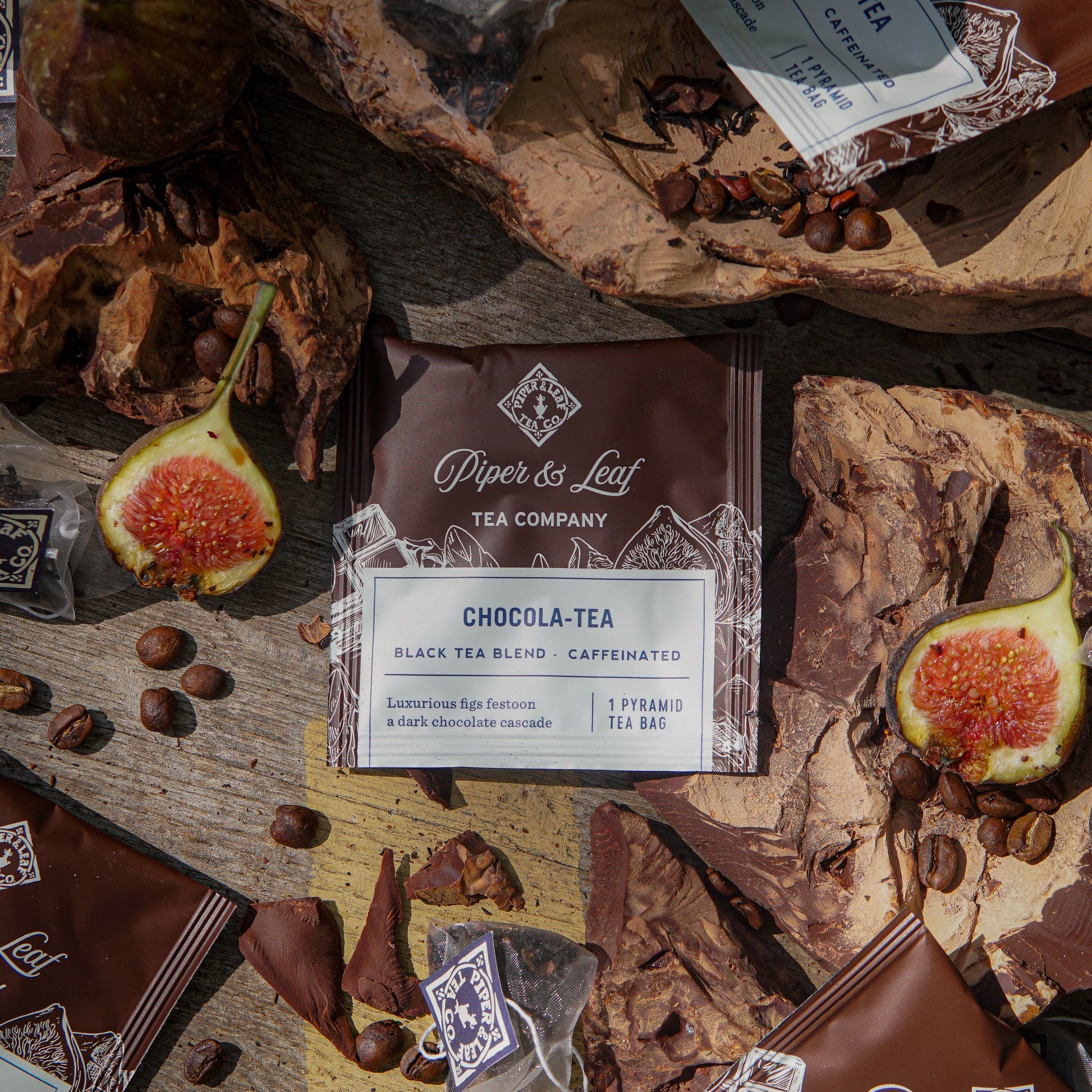 A box of Piper & Leaf Tea Co. Chocola-Tea Case of Individually Wrapped Tea Bags (50ct) is displayed on a wooden surface with fresh figs, cocoa pieces, whole coffee beans, and tea sachets arranged nearby.