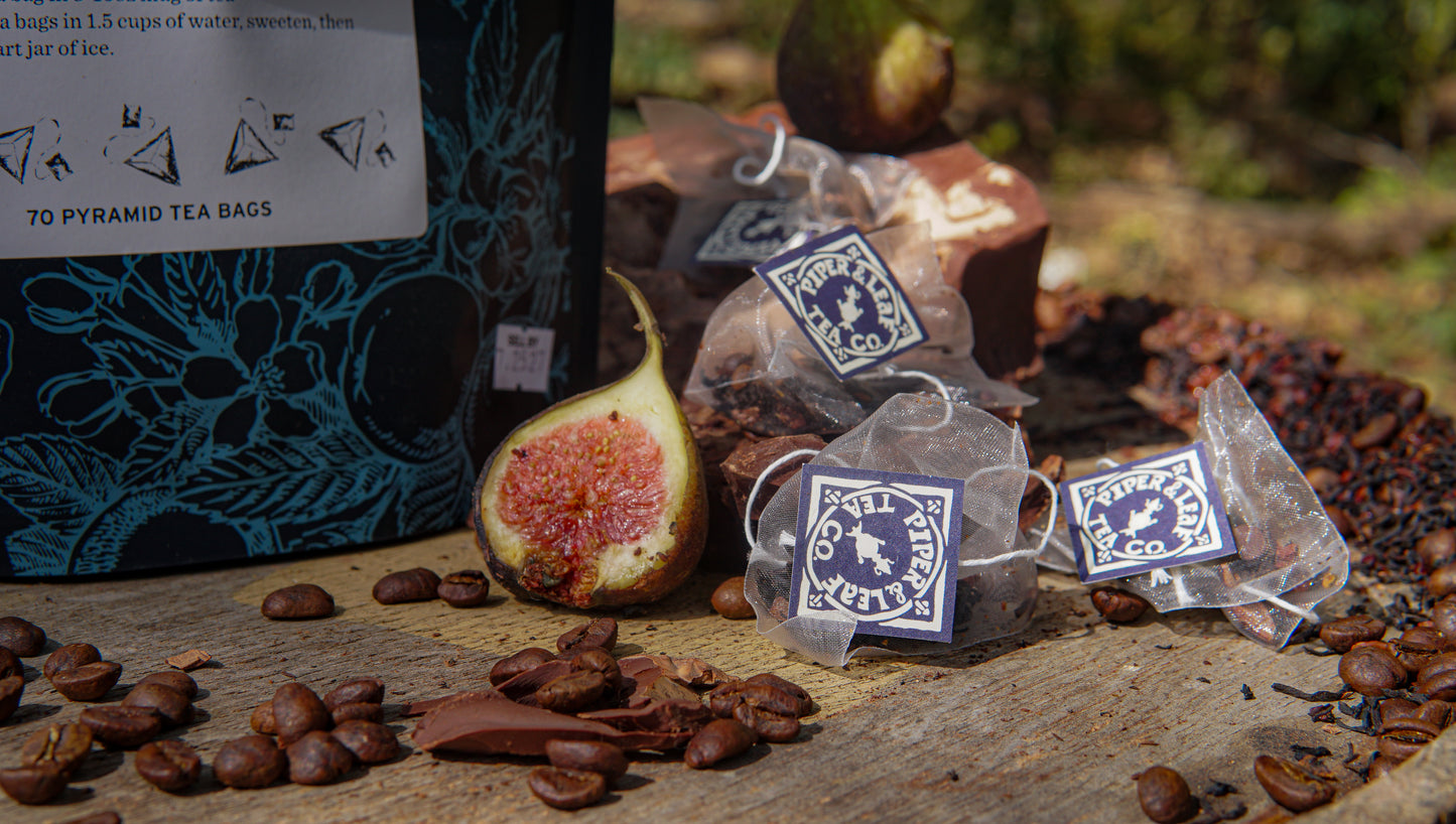 A close-up of loose coffee beans, a halved fig, chocolate pieces, and black tea sachets on a wooden surface next to a container labeled "Chocola-Tea Bulk Sachets - 70ct Tea Bags" by Piper & Leaf Tea Co.
