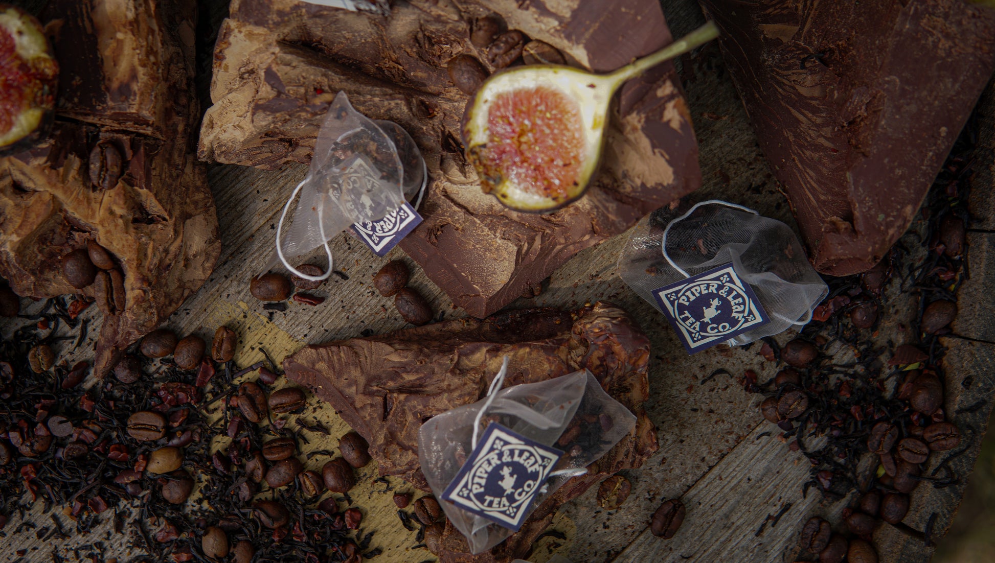 Three Piper & Leaf Tea Co. "Chocola-Tea Bulk Sachets" rest on chocolate chunks with coffee beans, loose tea leaves, and fig, evoking the rich flavors of dark chocolate and fig teas on a rustic wooden surface.