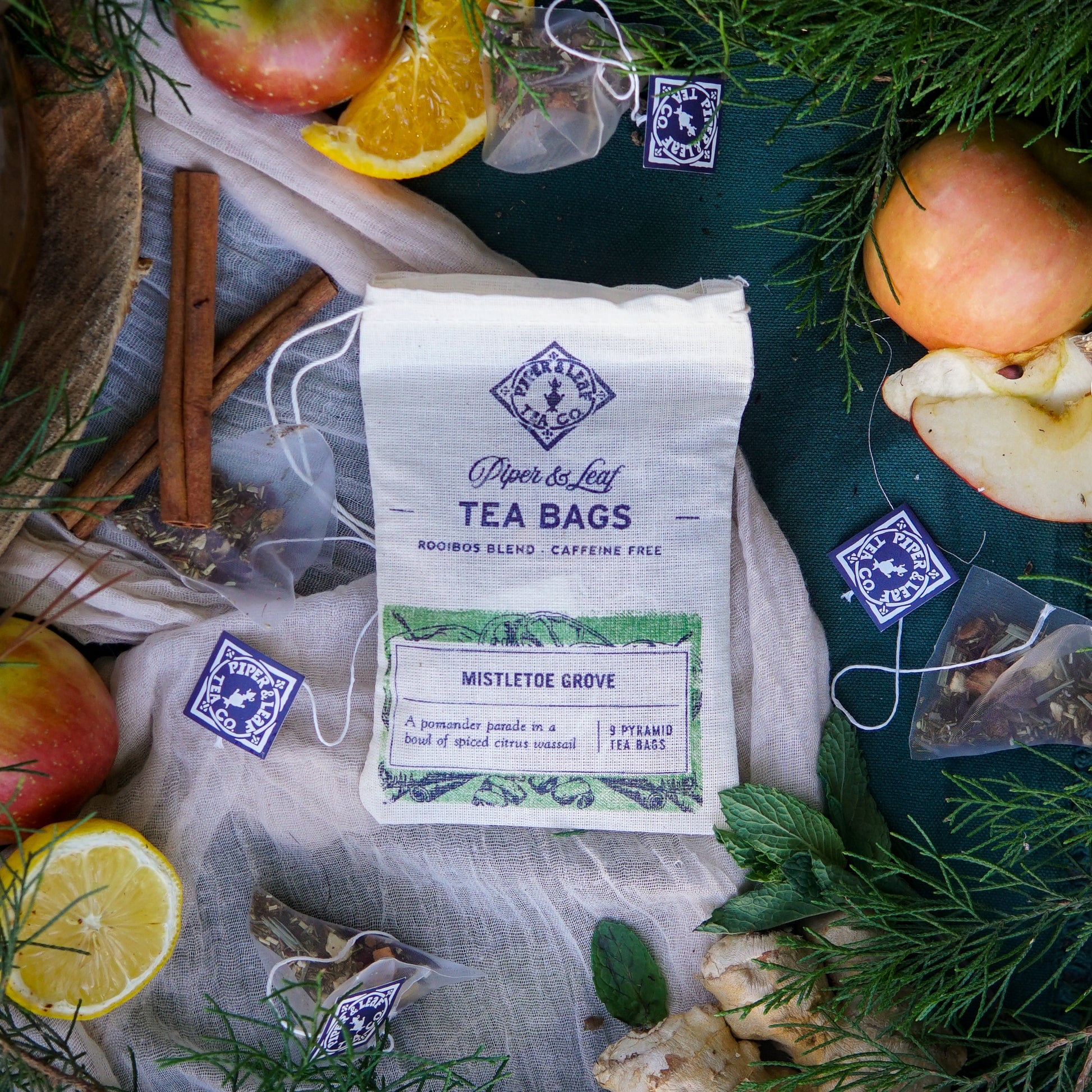 Piper & Leaf Tea Co.’s Mistletoe Grove 9ct Tea Bags in Muslin, a caffeine-free rooibos blend, is pictured with apples, cinnamon, ginger, herbs, and loose tea bags on a green cloth—ideal for holiday sipping.