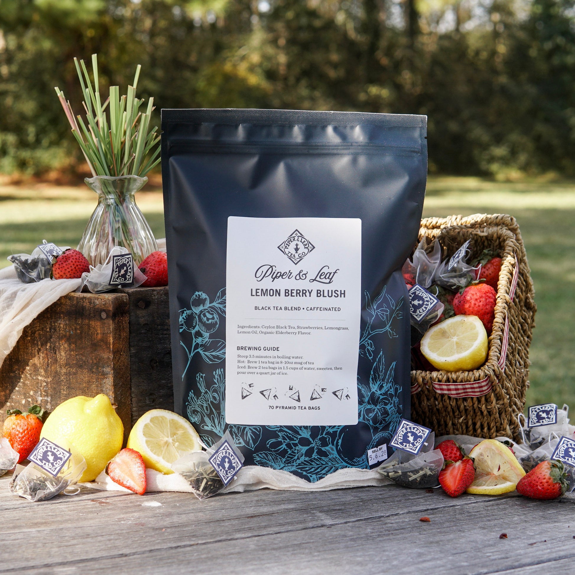 A box of Piper & Leaf Tea Co.'s Lemon Berry Blush Bulk Sachets (70ct tea bags) sits outdoors on a wooden surface, surrounded by lemons, strawberries, a wicker basket, and several tea bags.