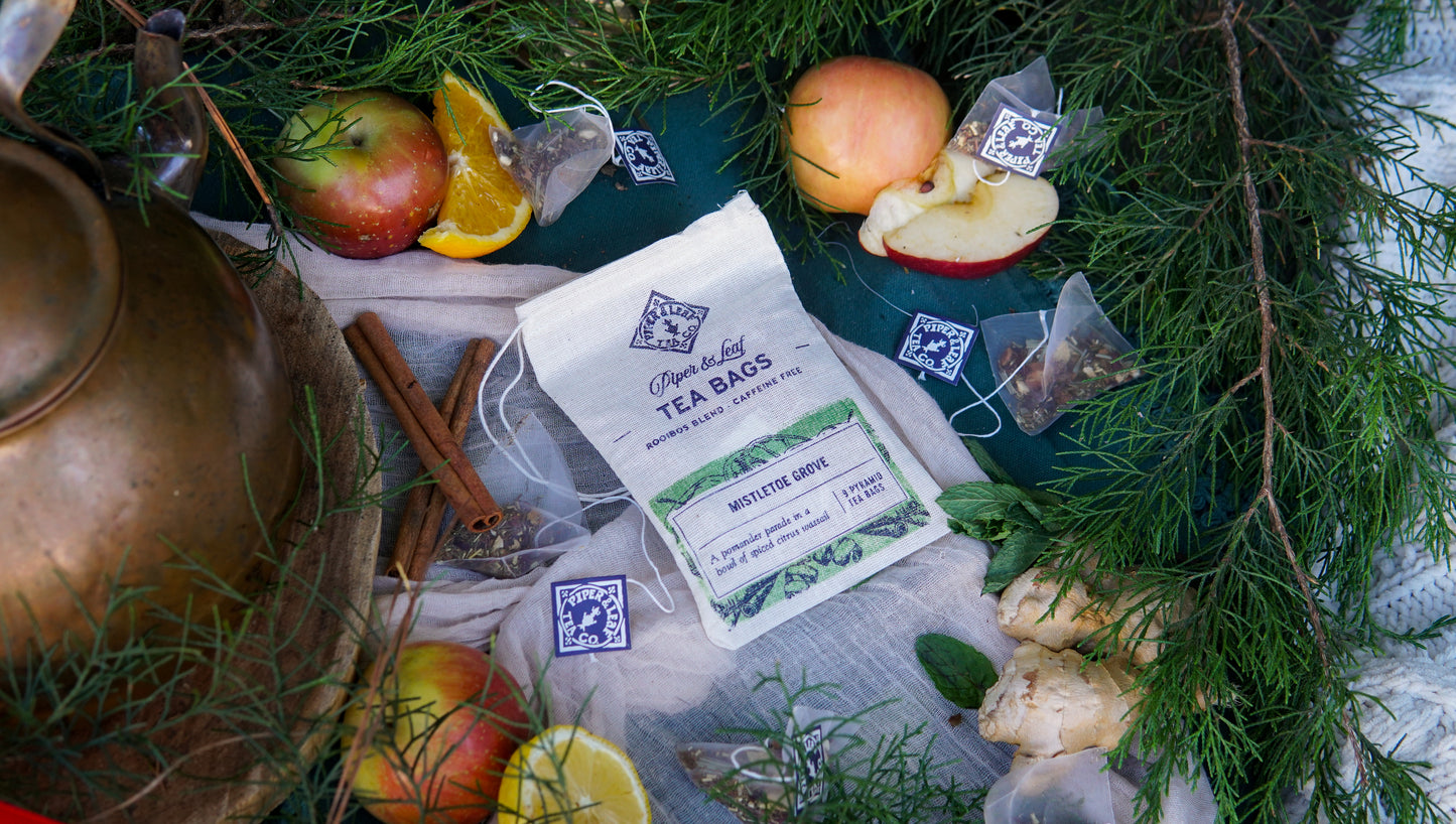 A packet of Piper & Leaf Tea Co.'s Mistletoe Grove 9ct Tea Bags in Muslin sits among apples, oranges, cinnamon sticks, ginger, and greenery—accompanied by loose tea bags and a brass kettle—for a festive caffeine-free treat.