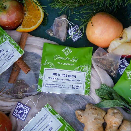 A case of 50 individually wrapped Mistletoe Grove tea bags by Piper & Leaf Tea Co., surrounded by ginger, apples, orange slices, cinnamon sticks, mint leaves, and loose tea bags.