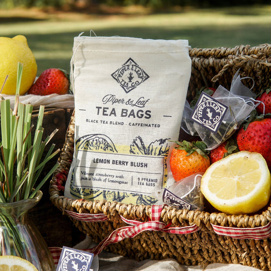 A bag of Piper & Leaf Tea Co. Lemon Berry Blush 9ct Tea Bags in Muslin rests in a woven basket with fresh strawberries, lemons, and tea sachets outdoors on a sunny day.
