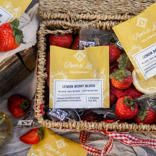 A woven tea gift basket featuring Piper & Leaf Tea Co.'s Lemon Berry Blush 50ct individually wrapped tea bags, fresh strawberries, sliced lemon, and extra tea bags and fruit arranged nearby.