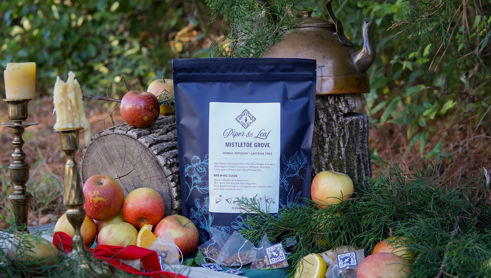 A bag of Piper & Leaf Tea Co.’s Mistletoe Grove Bulk Sachets (70ct) sits among apples, candles, greenery, and a brass kettle in a forest, evoking the warmth of a holiday tea ritual.