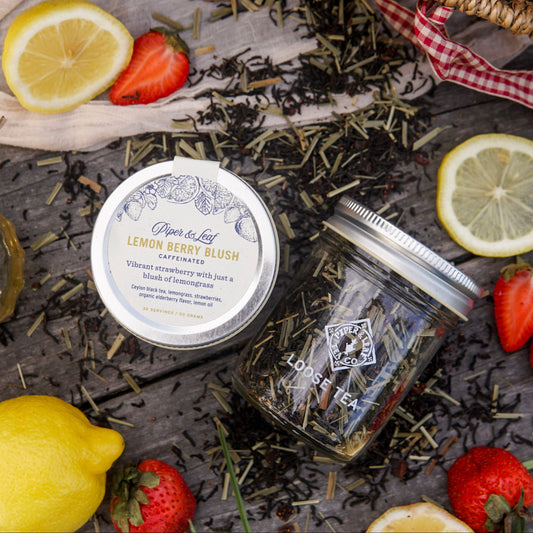 Two Piper & Leaf Tea Co. Lemon Berry Blush Glass Jars of Loose Leaf Tea (30 servings each) on wood, surrounded by strawberries, lemons, and lemongrass—ideal for lovers of strawberry and lemongrass tea.