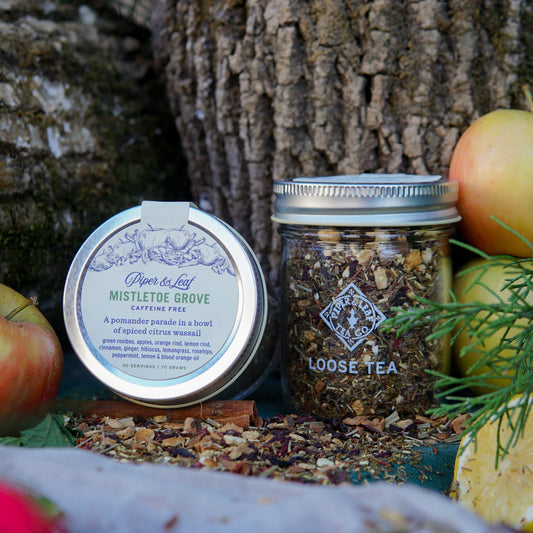 A glass jar of Piper & Leaf Tea Co.'s Mistletoe Grove Loose Leaf Tea (30 Servings) sits outdoors among apples and greenery, inviting you to enjoy this caffeine-free green rooibos blend in nature’s embrace.