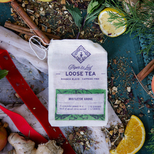 A Piper & Leaf Tea Co. Mistletoe Grove Muslin Bag of Loose Leaf Tea (15 Servings) sits on fabric, surrounded by festive ingredients like loose leaf tea, orange slices, and cinnamon sticks—a caffeine-free holiday blend for cozy moments.