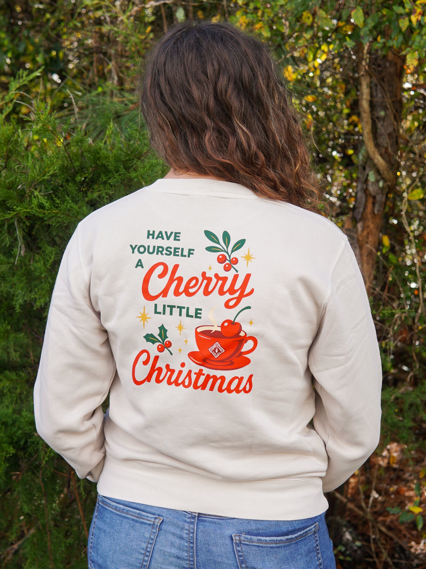 A person outdoors wears the Piper & Leaf Tea Co. "A Cherry Little Christmas Shirt," featuring festive graphics and the phrase "Have yourself a cherry little Christmas" on the back—a perfect holiday gift for any tea lover.