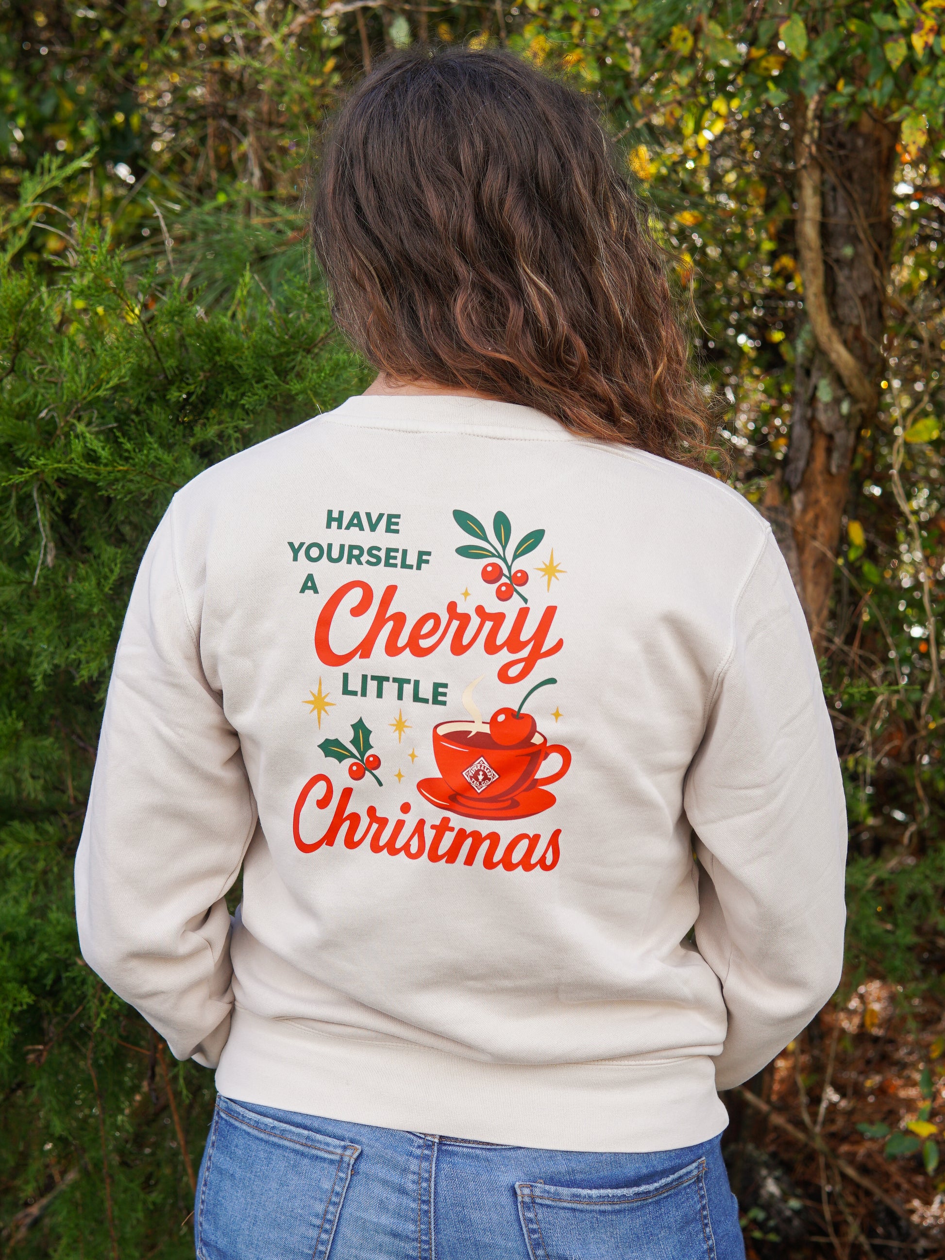 A person outdoors wears the Piper & Leaf Tea Co. "A Cherry Little Christmas Shirt," featuring festive graphics and the phrase "Have yourself a cherry little Christmas" on the back—a perfect holiday gift for any tea lover.