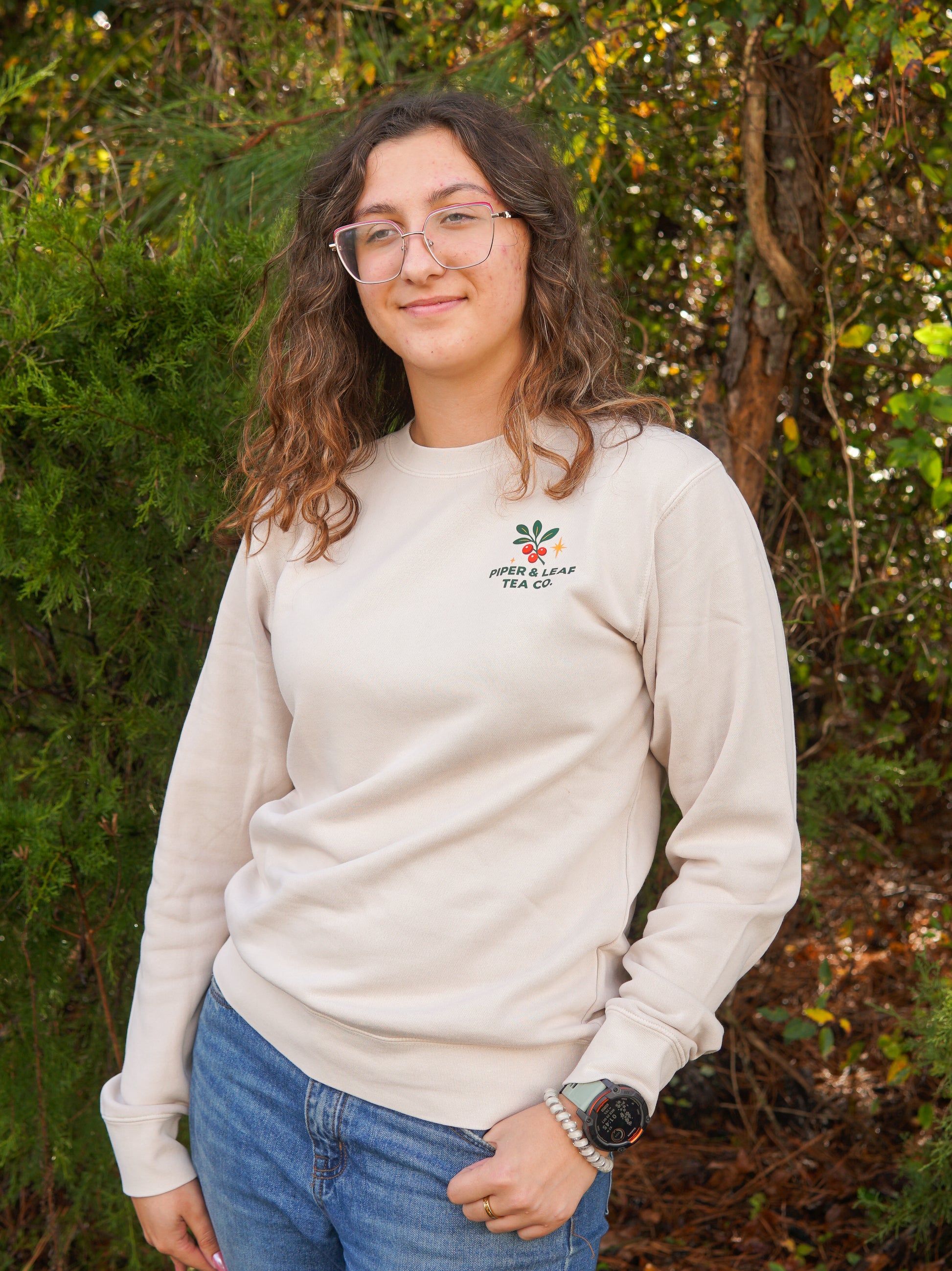 A person with wavy brown hair and glasses stands outdoors in front of greenery, wearing Piper & Leaf Tea Co.'s A Cherry Little Christmas Shirt and blue jeans—a perfect holiday gift for tea lovers.