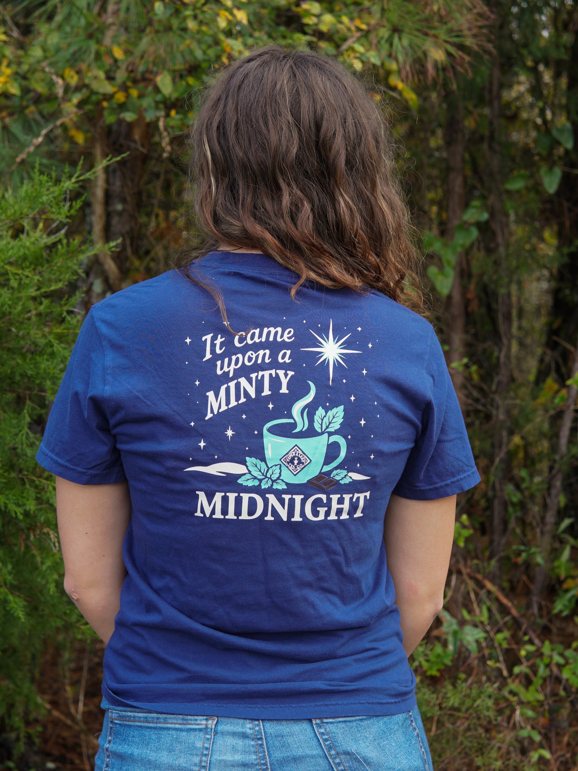 A person with long brown hair stands outdoors wearing the Piper & Leaf Tea Co.'s "It Came Upon a Minty Midnight" T-shirt, featuring a steaming cup and stars graphic.