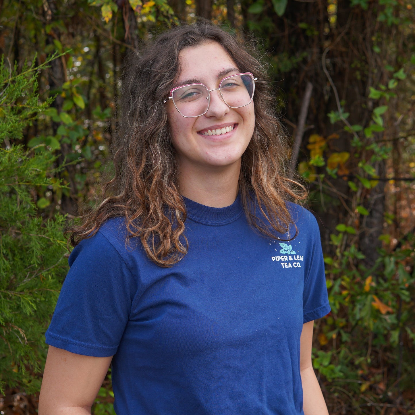 A person with long wavy hair and glasses, smiling at the camera, stands outdoors in front of greenery while wearing a blue "It Came Upon a Minty Midnight" t-shirt from Piper & Leaf Tea Co.