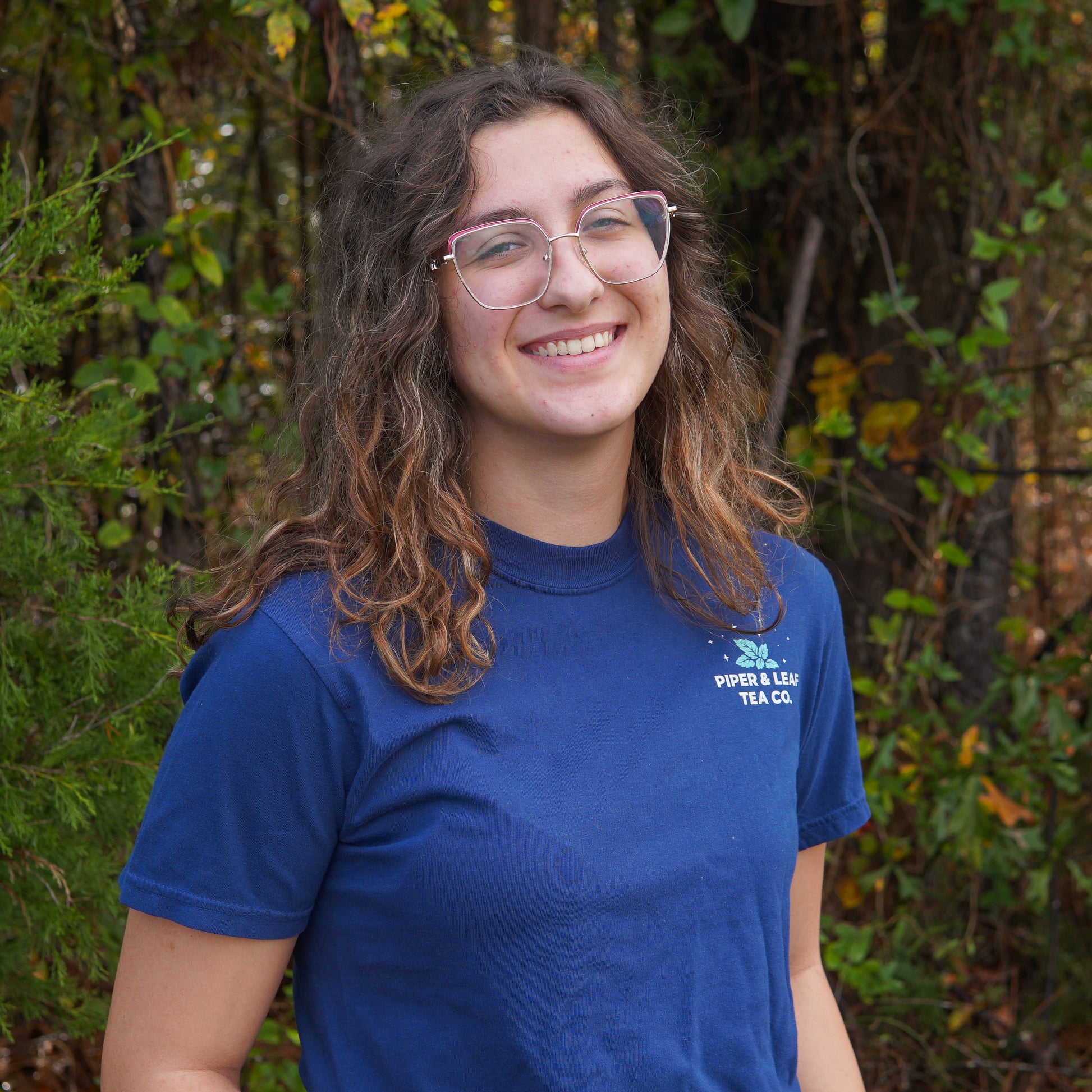 A person with long wavy hair and glasses, smiling at the camera, stands outdoors in front of greenery while wearing a blue "It Came Upon a Minty Midnight" t-shirt from Piper & Leaf Tea Co.