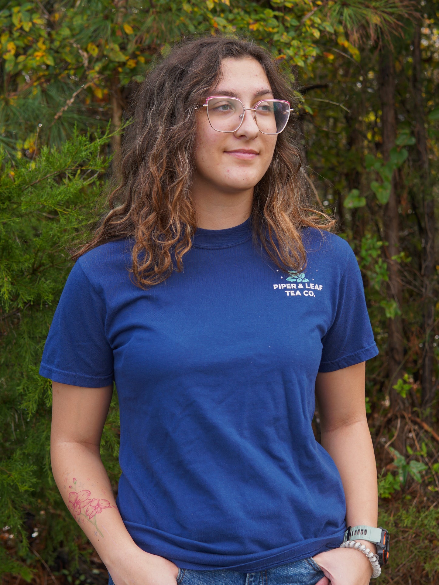 A person with wavy brown hair and glasses stands outdoors in a blue "It Came Upon a Minty Midnight" T-Shirt by Piper & Leaf Tea Co., surrounded by greenery.