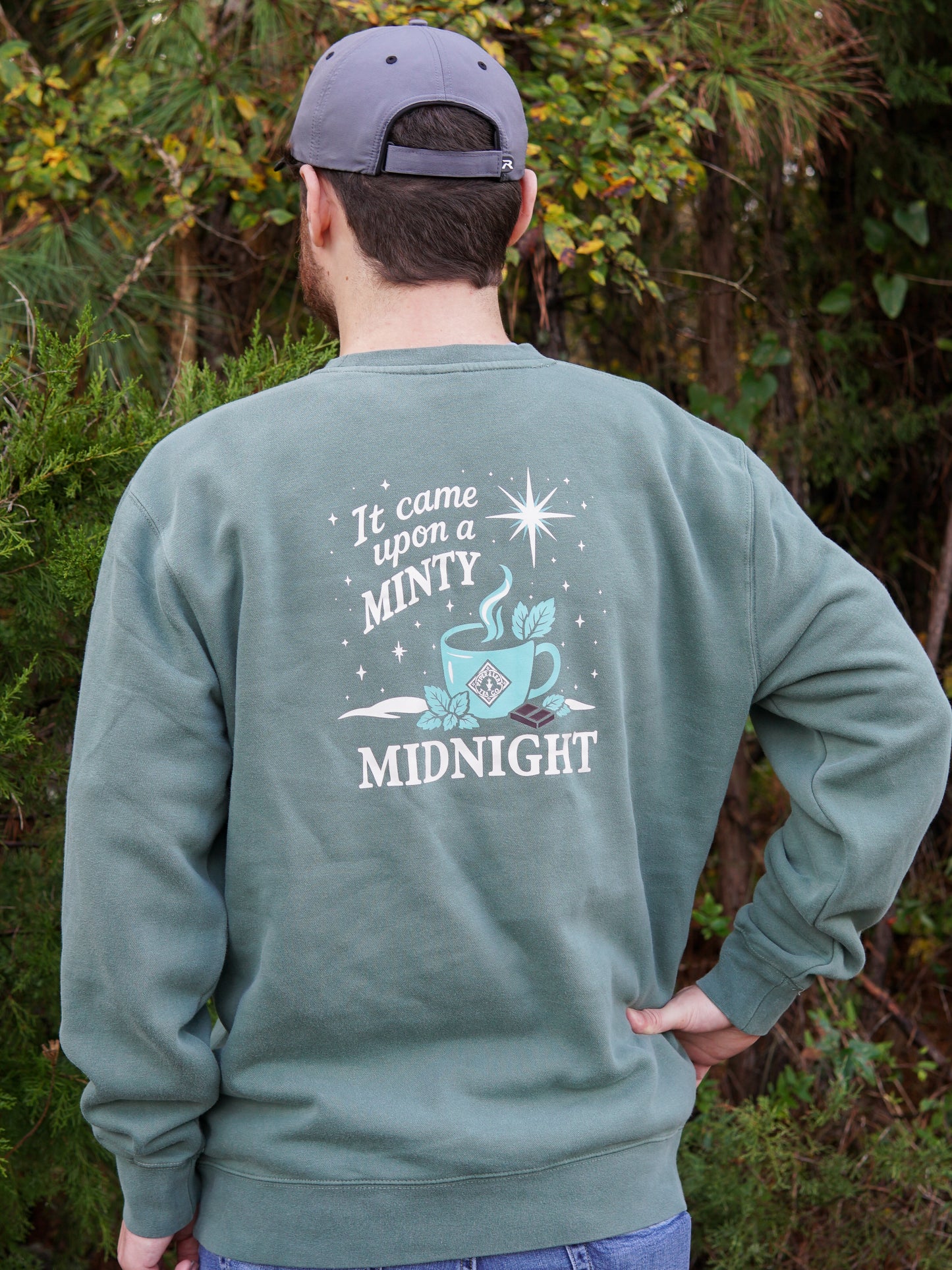 A person wears the cozy Piper and Leaf Tea Co. "It Came Upon a Minty Midnight Sweatshirt," featuring a teacup graphic and text, while outdoors facing away. Perfect for relaxing, caffeine-free evenings.