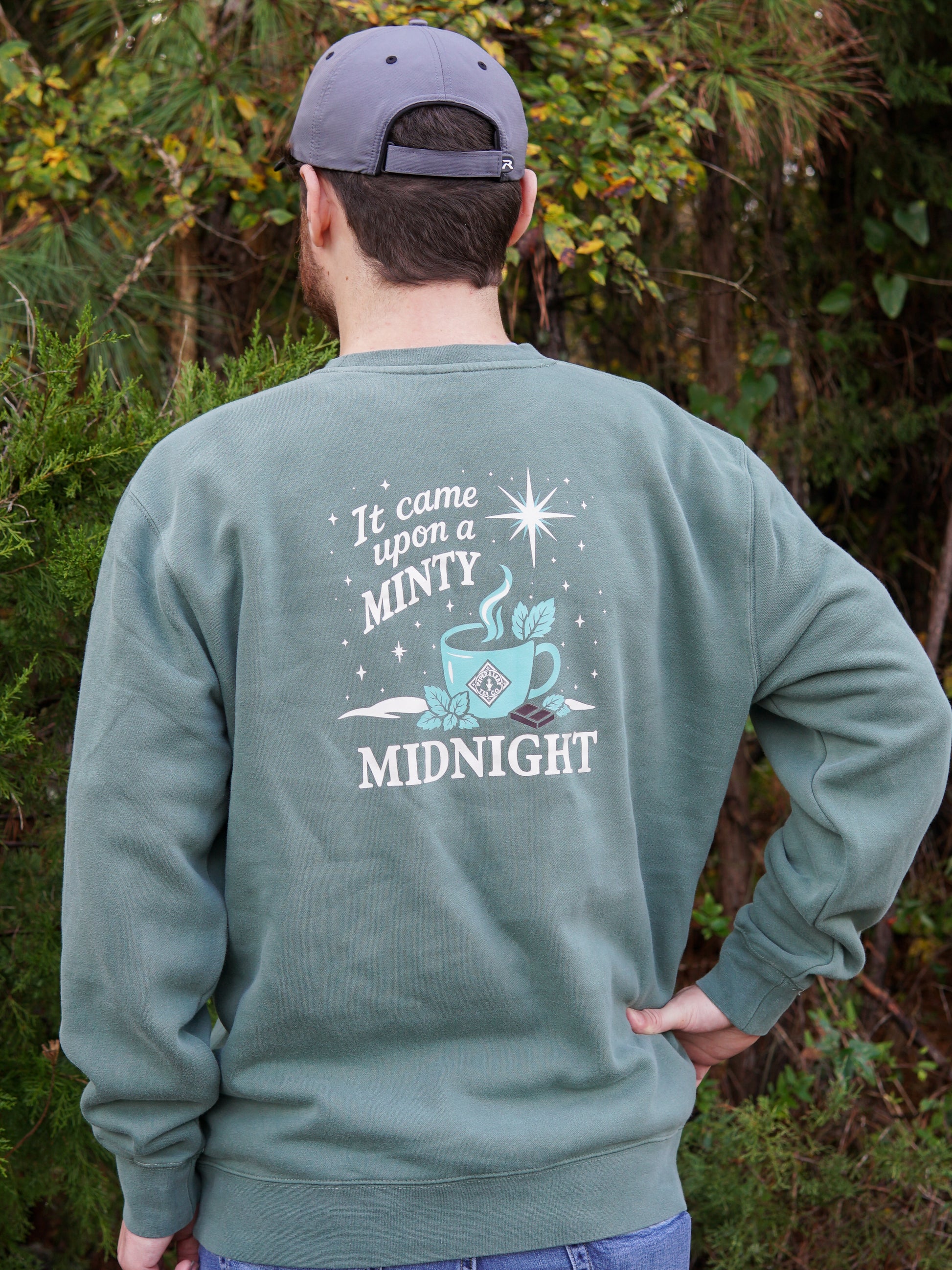 A person wears the cozy Piper and Leaf Tea Co. "It Came Upon a Minty Midnight Sweatshirt," featuring a teacup graphic and text, while outdoors facing away. Perfect for relaxing, caffeine-free evenings.
