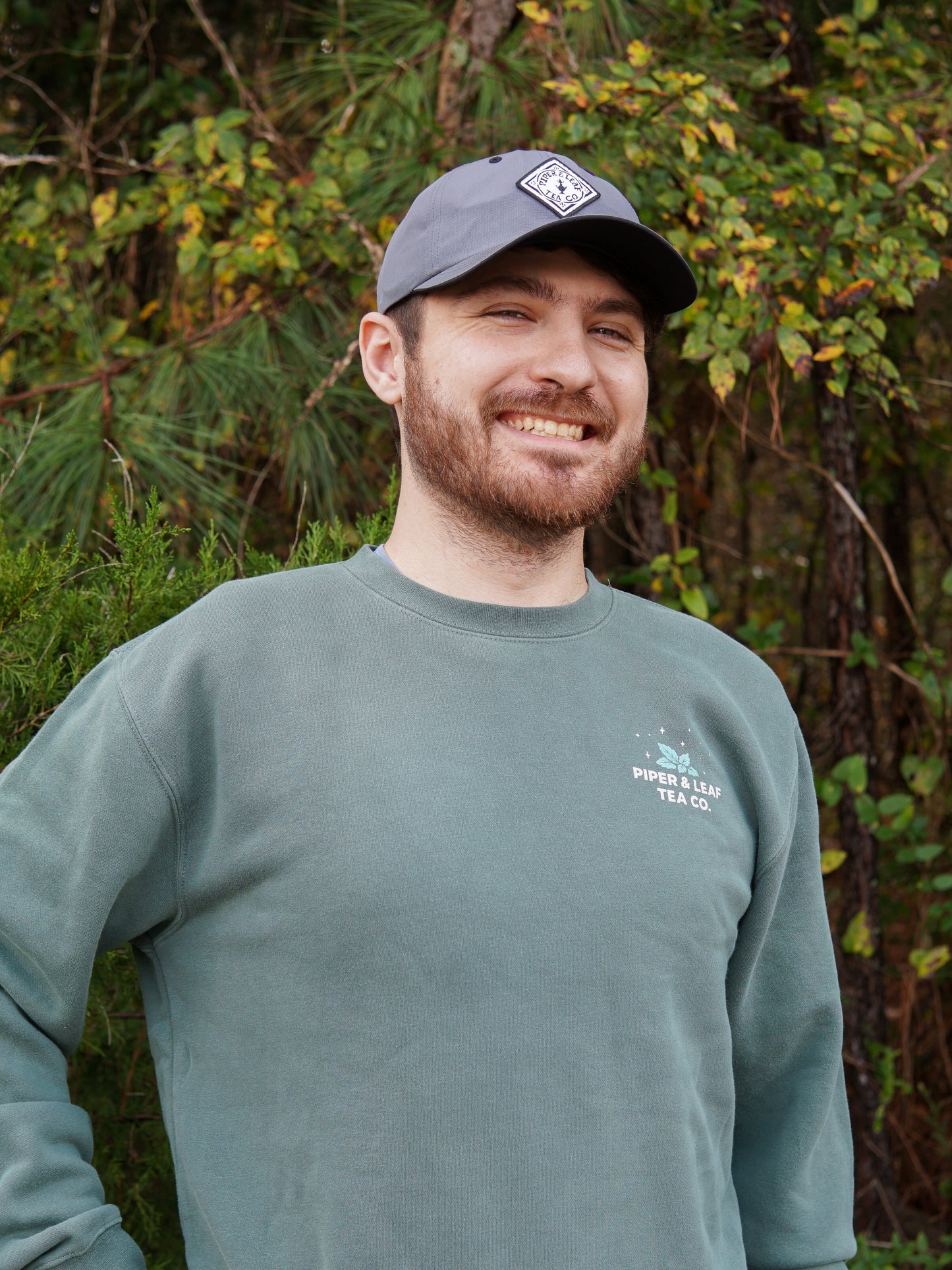 A bearded man in a “Piper and Leaf Tea Co.” It Came Upon a Minty Midnight Sweatshirt and gray cap smiles outdoors by leafy trees, perhaps savoring the fresh vibe of this cozy, caffeine-free inspired look.