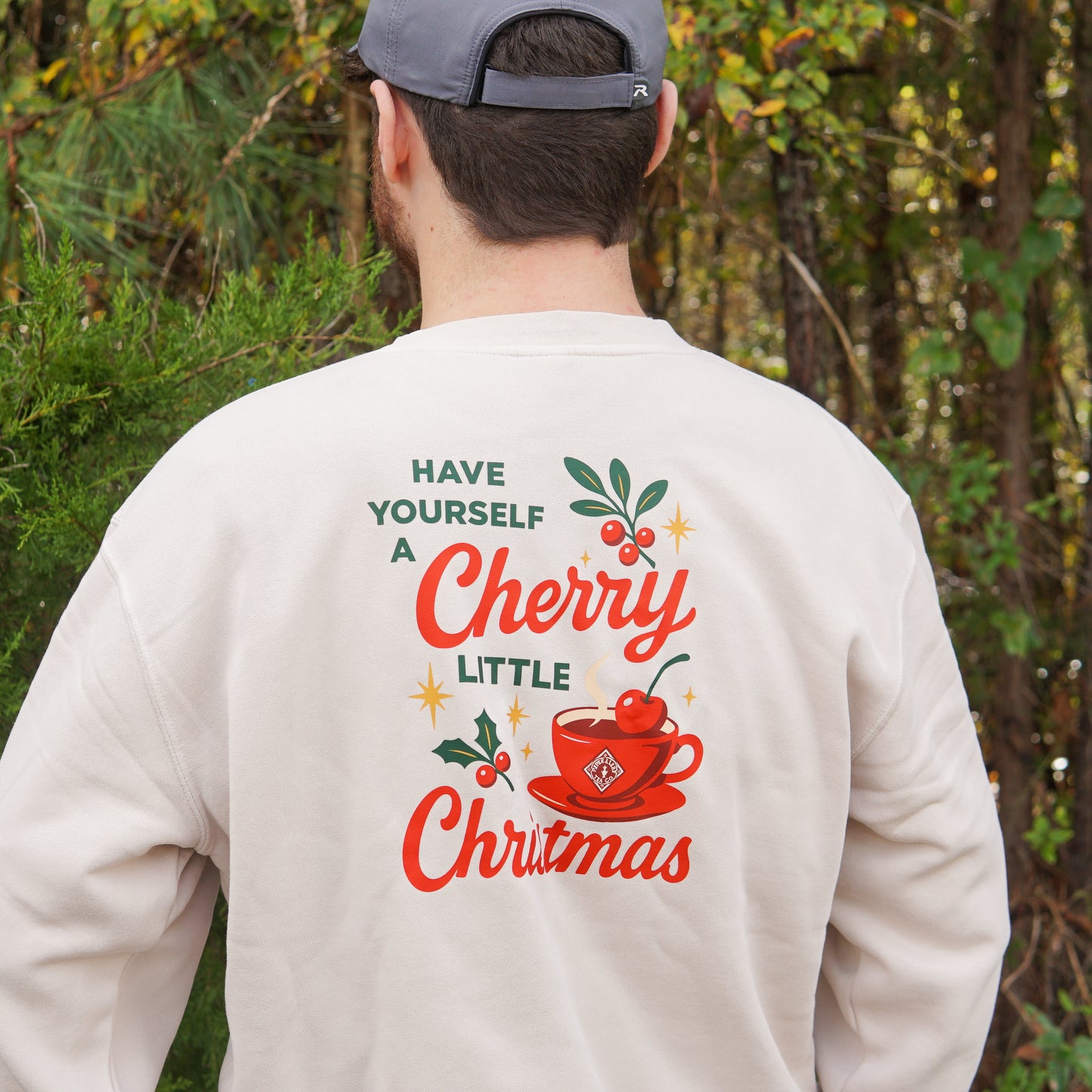 A person wearing the Piper & Leaf Tea Co. "A Cherry Little Christmas Shirt" featuring a red teacup stands outdoors by trees—the ideal gift for tea lovers.