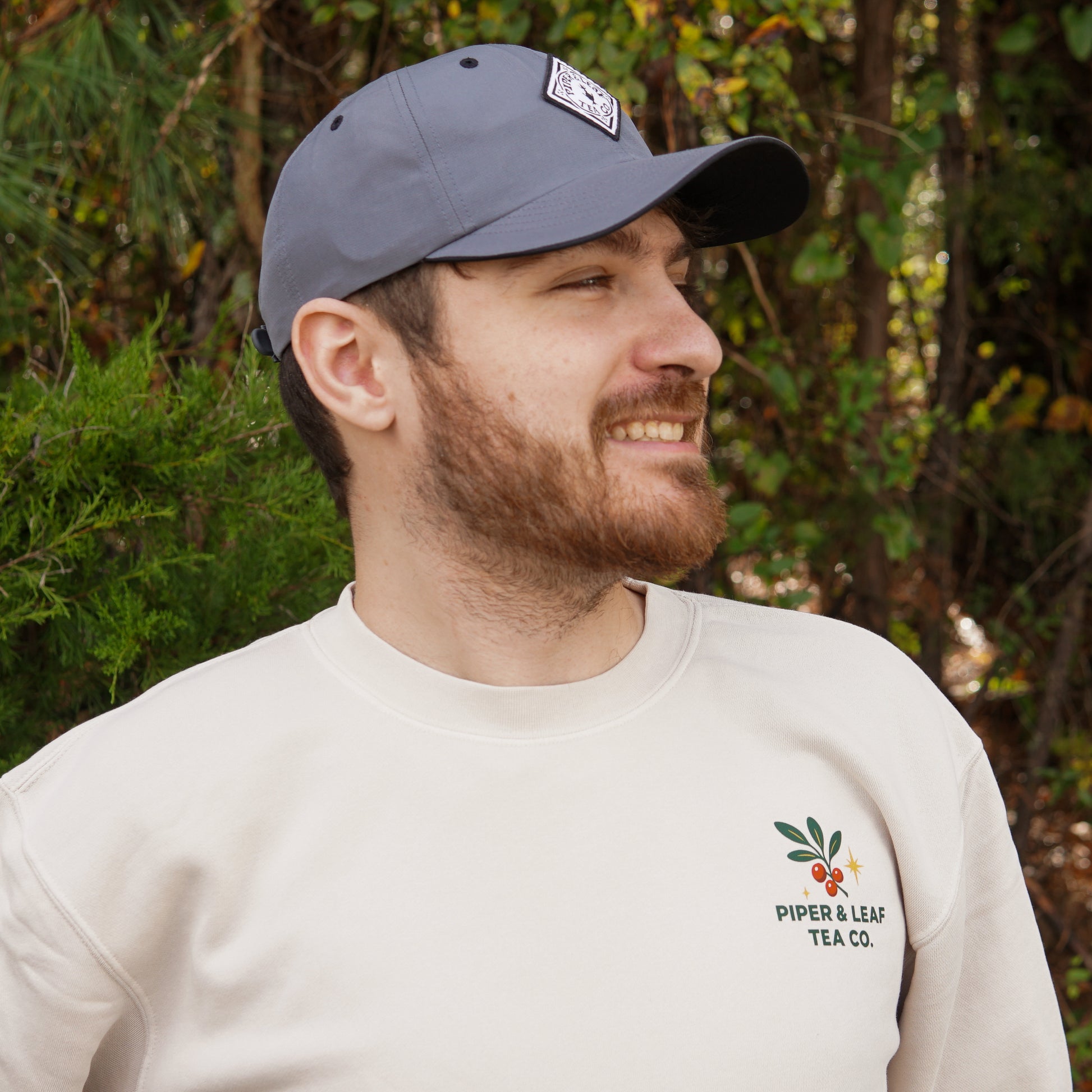 A bearded man in a gray cap and light "A Cherry Little Christmas Shirt" by Piper & Leaf Tea Co. stands smiling outdoors amid green foliage—a festive look for tea lovers.
