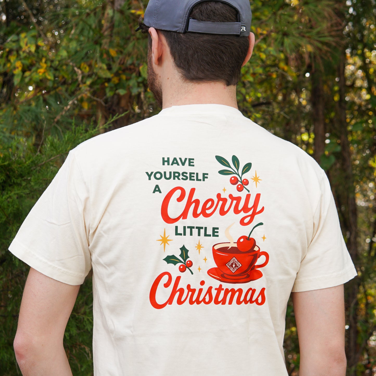 A man in a Piper & Leaf Tea Co. "A Cherry Little Christmas Shirt" with a cherry-topped teacup stands outdoors by trees—an ideal holiday tea lover gift.