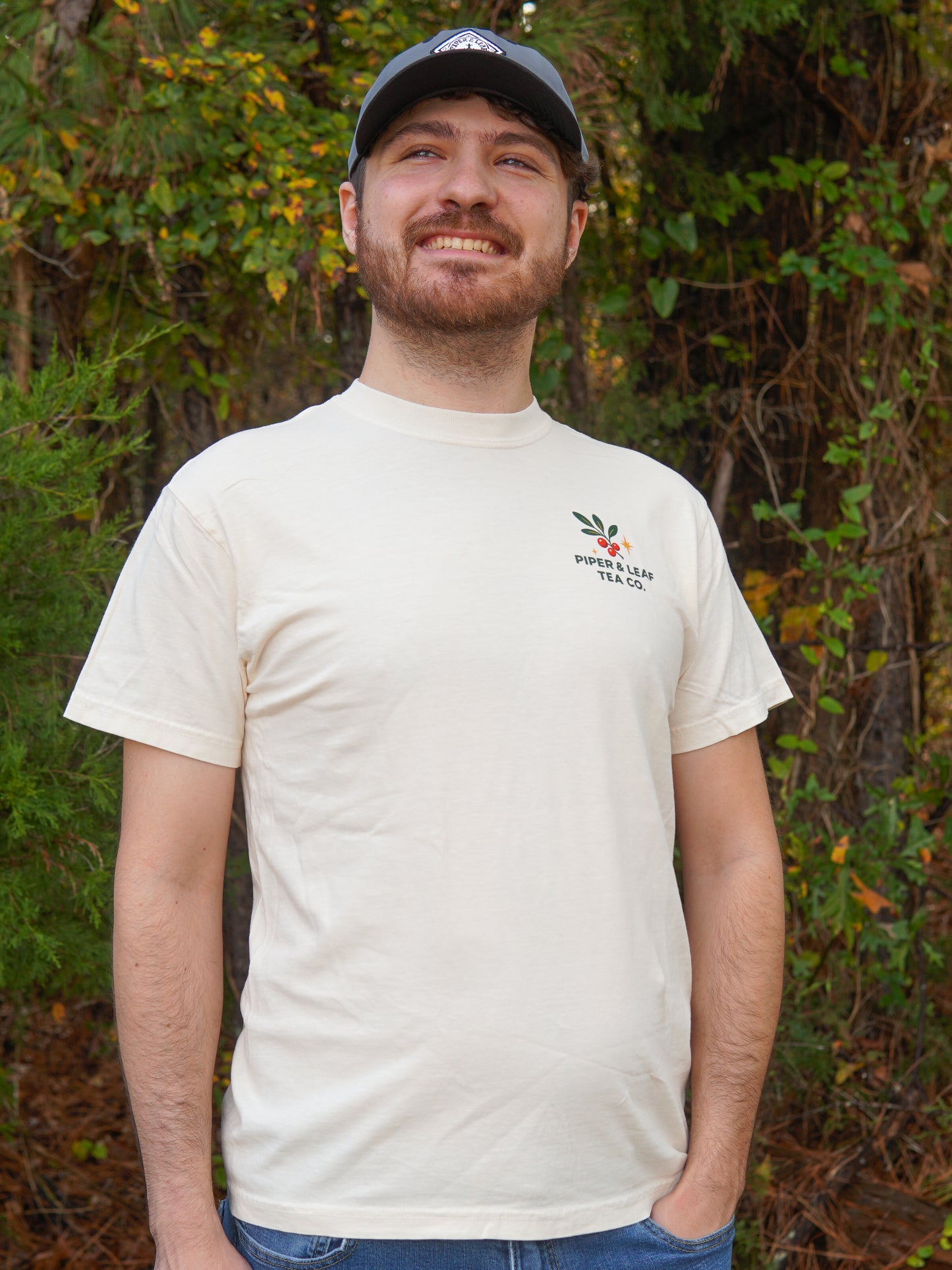 A bearded man in a black cap and a cream "A Cherry Little Christmas Shirt" by Piper & Leaf Tea Co. smiles outdoors before green foliage—the perfect gift for tea lovers.