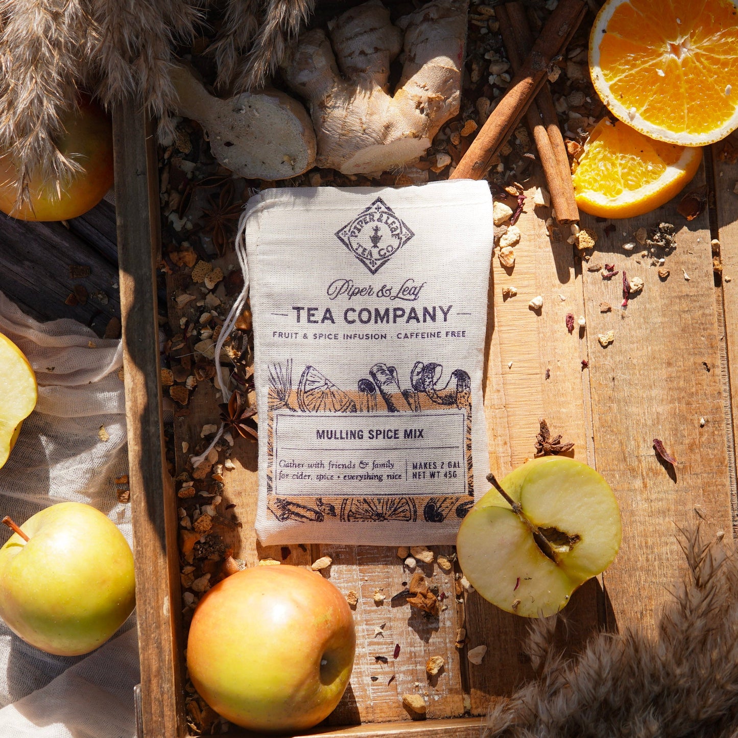 A bag of Piper & Leaf Tea Co. Mulling Spice Mix rests in a wooden tray, surrounded by apples, orange slices, spices, and ginger root—ideal for making spiced cider at holiday gatherings.