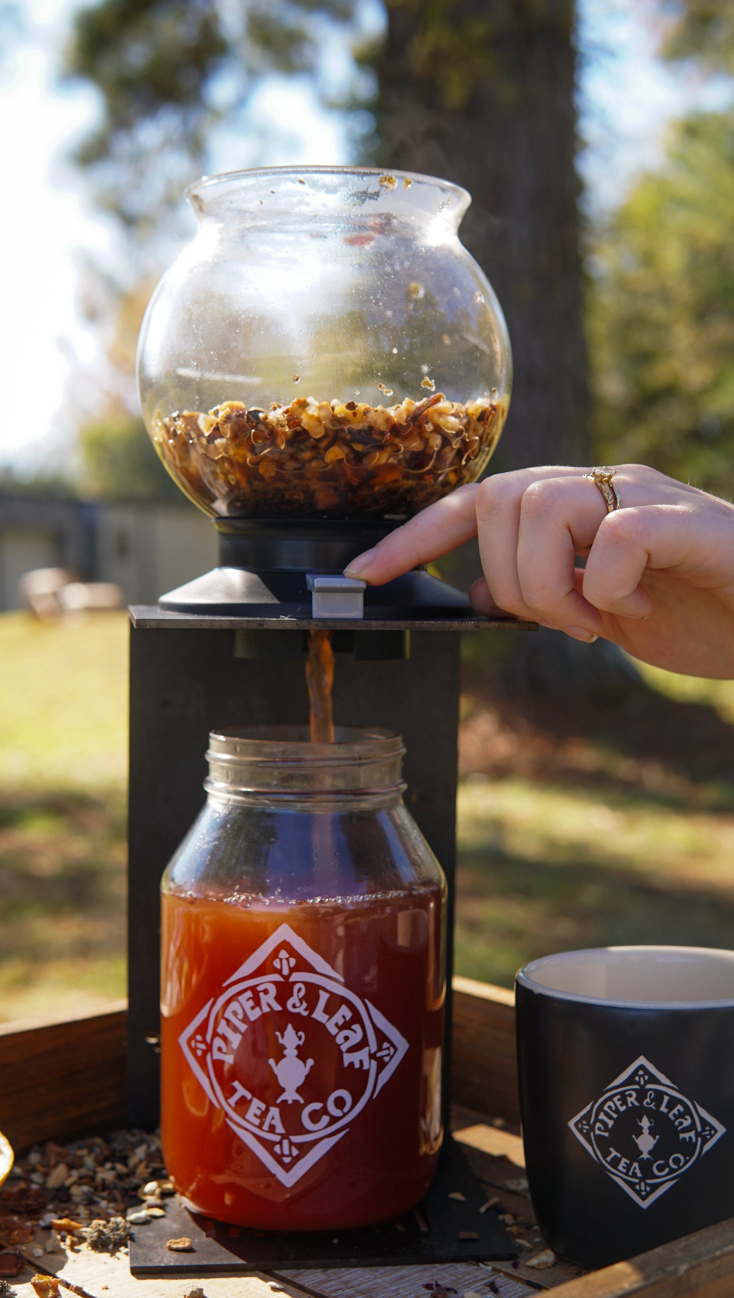 Someone pours tea from a glass infuser into a "Piper & Leaf Tea Co." jar at a holiday party. Nearby, a black Piper & Leaf mug suggests serving their Mulling Spice Mix—ideal for festive, spiced drinks outdoors.