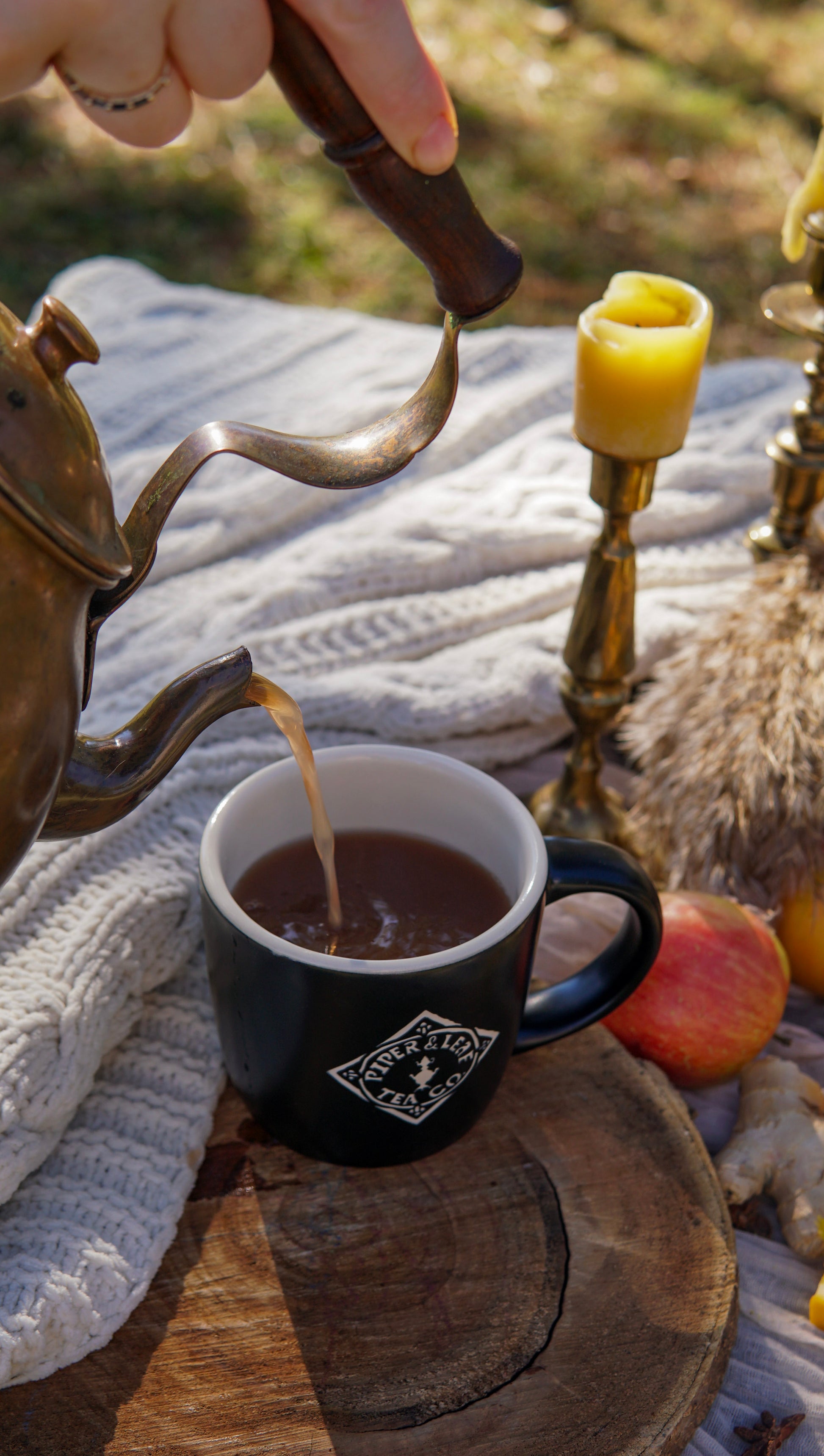 A hand pours tea made with Piper & Leaf Tea Co.'s Mulling Spice Mix from a brass teapot into a black mug, surrounded by a white knit blanket, yellow candles, and an apple on a cozy picnic setup.