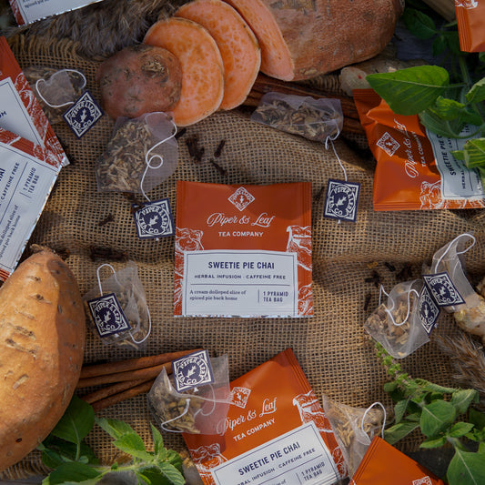 Piper & Leaf Tea Co. Sweetie Pie Chai Case of Tea Bag Envelopes (50ct) displayed with bread, sweet potato, cinnamon sticks, and green leaves on burlap. This caffeine-free chai blend delivers cozy, comforting flavors.