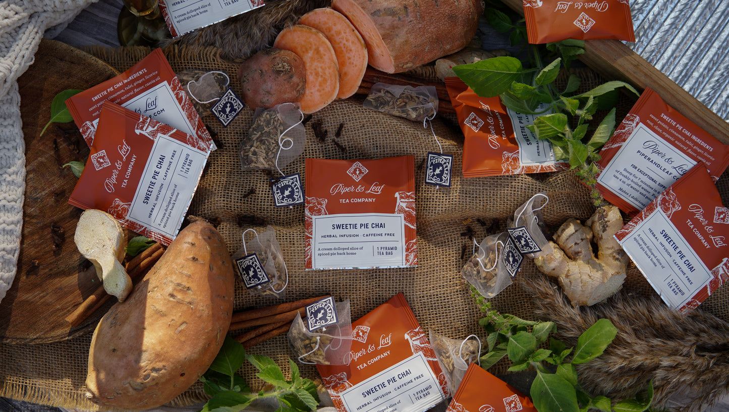 A case of 50 Sweetie Pie Chai Tea Bag Envelopes by Piper & Leaf Tea Co. displayed with sweet potatoes, ginger, and fresh herbs on a wooden surface with a textured cloth.