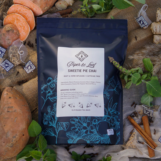 A package of Piper & Leaf Tea Co. Sweetie Pie Chai Bulk Sachets (70ct Tea Bags), a caffeine-free spiced herbal tea, is displayed with sweet potatoes, cinnamon sticks, ginger, tea bags, and fresh herbs on a rustic surface.