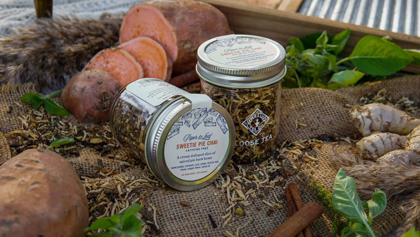 Two jars of Piper & Leaf Tea Co.'s Sweetie Pie Chai Glass Jar of Loose Leaf Tea (30 servings) rest on burlap with sweet potatoes, ginger, cinnamon sticks, and greens—perfect for a cozy cup of this caffeine-free blend.