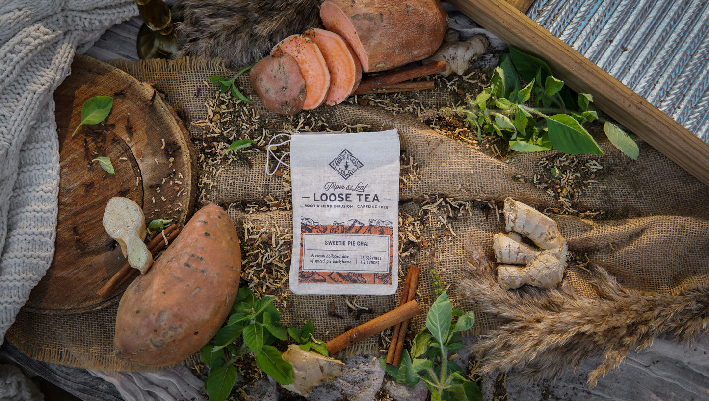 A Piper & Leaf Tea Co. Sweetie Pie Chai Muslin Bag of Loose Leaf Tea (15 servings) is set amid sweet potatoes, ginger, cinnamon sticks, leaves, and rustic fabric on a textured surface, evoking the cozy feel of candied sweet potato tea.