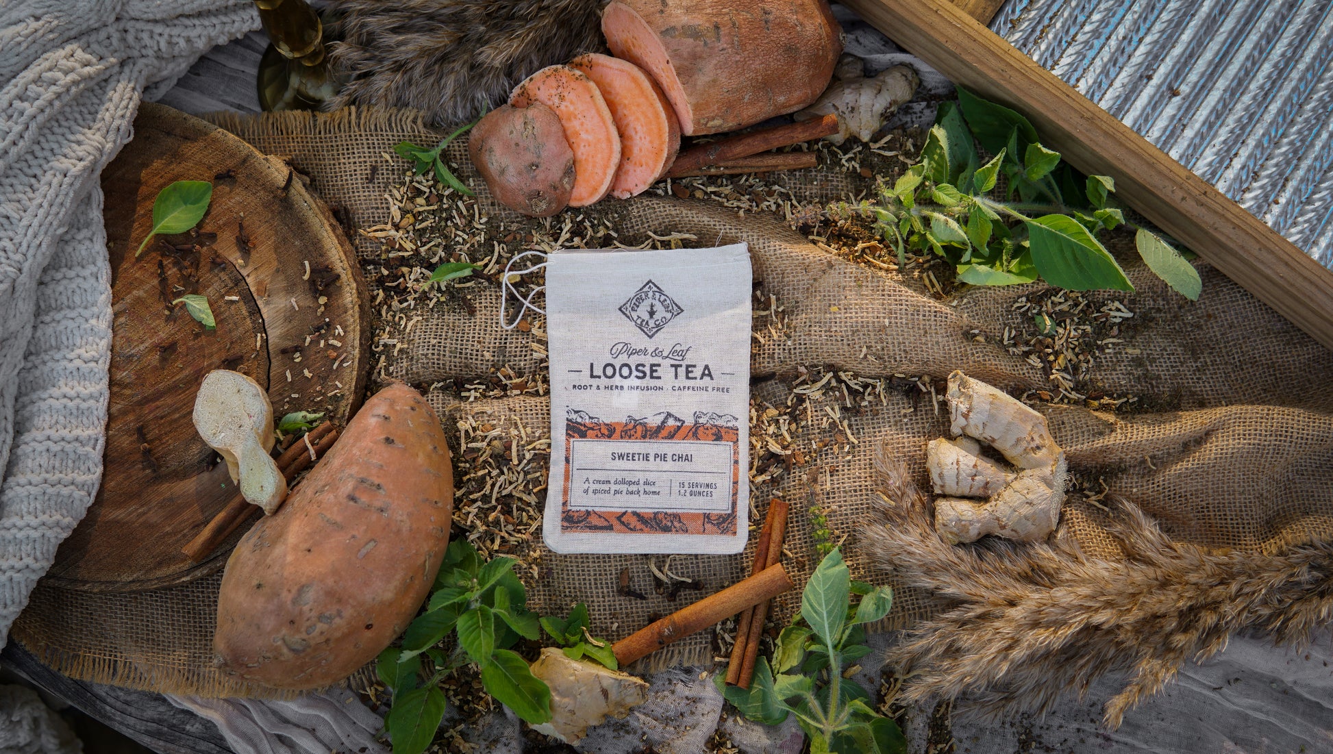 A Piper & Leaf Tea Co. Sweetie Pie Chai Muslin Bag of Loose Leaf Tea (15 servings) is set amid sweet potatoes, ginger, cinnamon sticks, leaves, and rustic fabric on a textured surface, evoking the cozy feel of candied sweet potato tea.