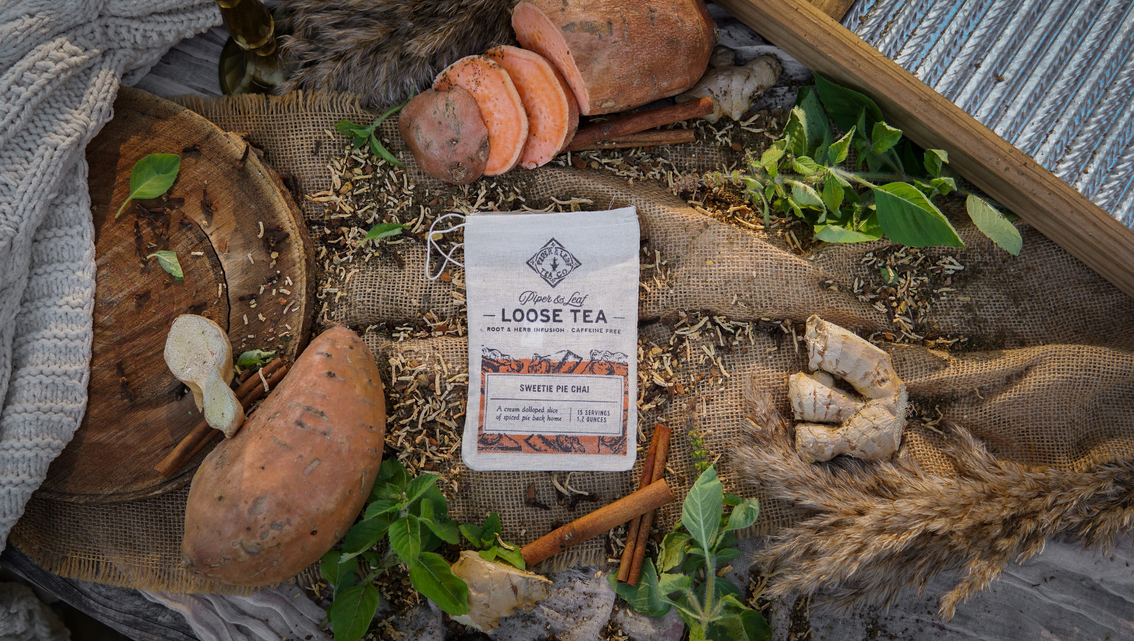 A Piper & Leaf Tea Co. Sweetie Pie Chai Muslin Bag of Loose Leaf Tea (15 servings) is set amid sweet potatoes, ginger, cinnamon sticks, leaves, and rustic fabric on a textured surface, evoking the cozy feel of candied sweet potato tea.