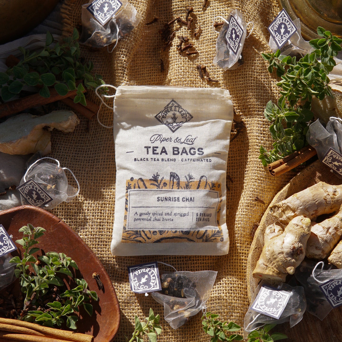 A muslin pouch labeled "Sunrise Chai 9ct Tea Bags" by Piper & Leaf Tea Co. sits on burlap, surrounded by chai tea bags, ginger roots, and fresh herbs.