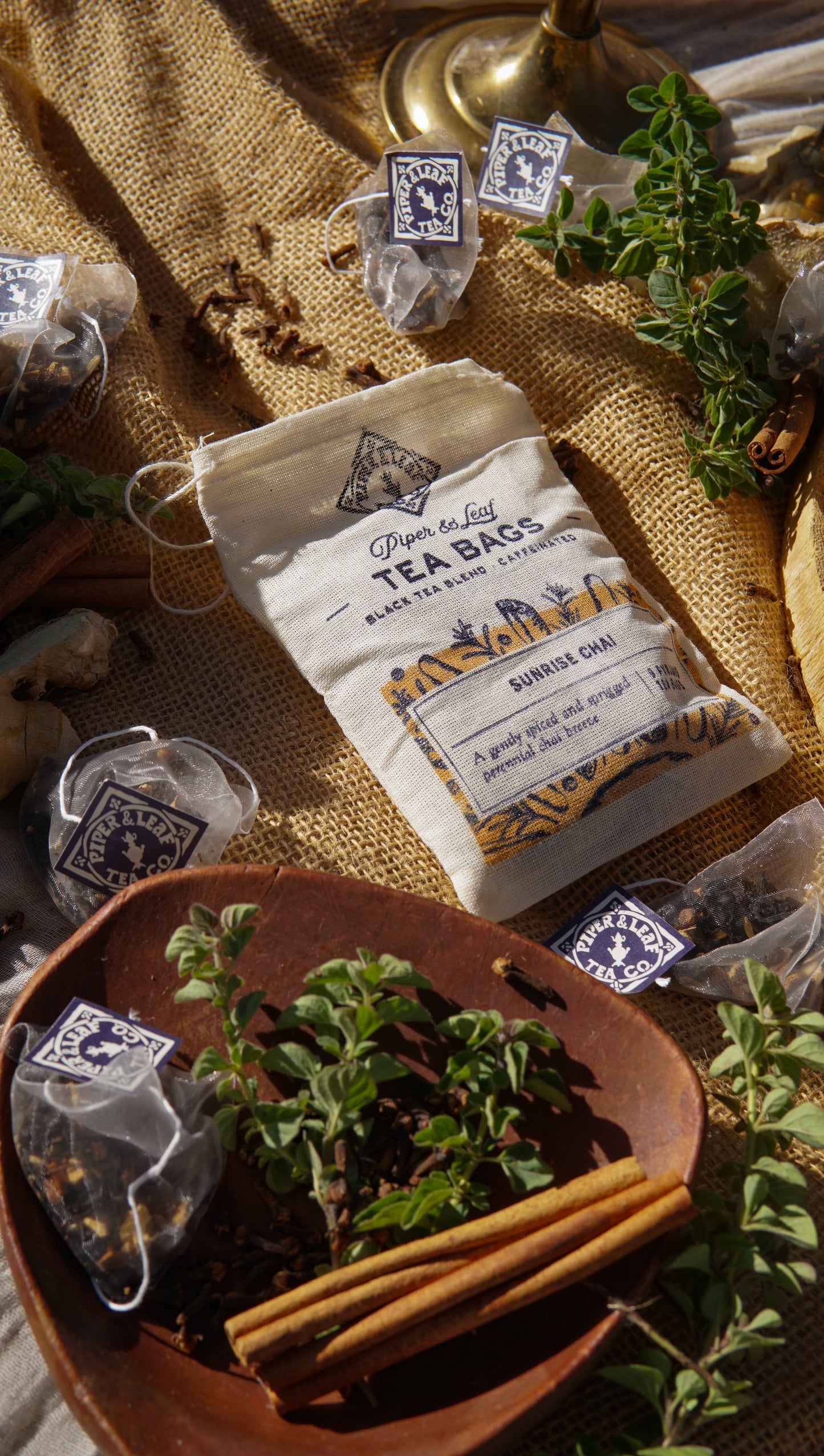 A muslin bag labeled "Sunrise Chai 9ct Tea Bags" by Piper & Leaf Tea Co. rests on burlap, surrounded by tea bags, herbs, cinnamon sticks, and garlic—ideal for fans of spiced chai or Nilgiri black tea blends.