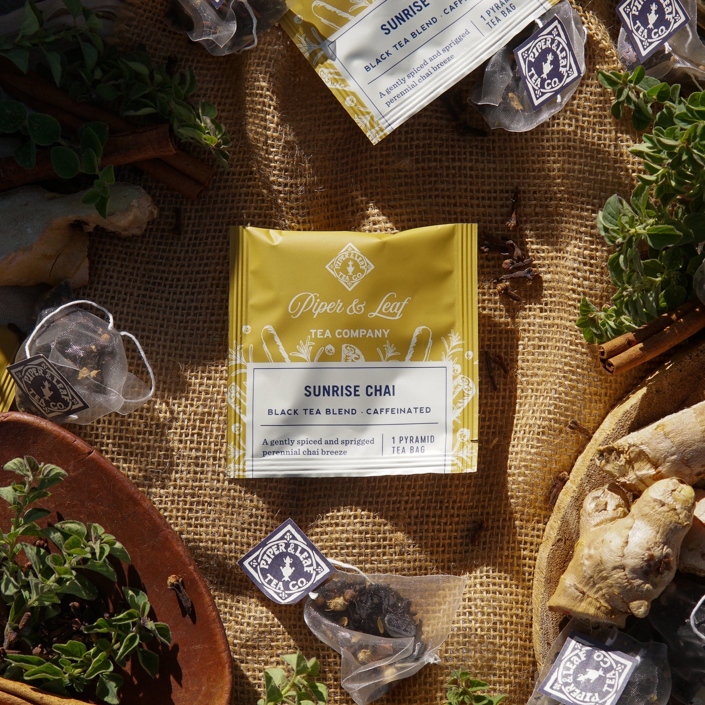 A yellow Sunrise Chai Case of Individually Wrapped Tea Bags (50ct) by Piper & Leaf Tea Co. is displayed on burlap, surrounded by single tea sachets, loose herbs, ginger root, and branded labels.