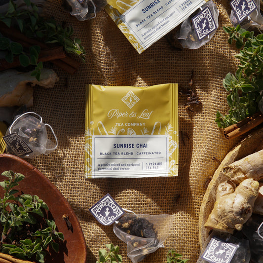 A yellow Sunrise Chai Case of Individually Wrapped Tea Bags (50ct) by Piper & Leaf Tea Co. is displayed on burlap, surrounded by single tea sachets, loose herbs, ginger root, and branded labels.