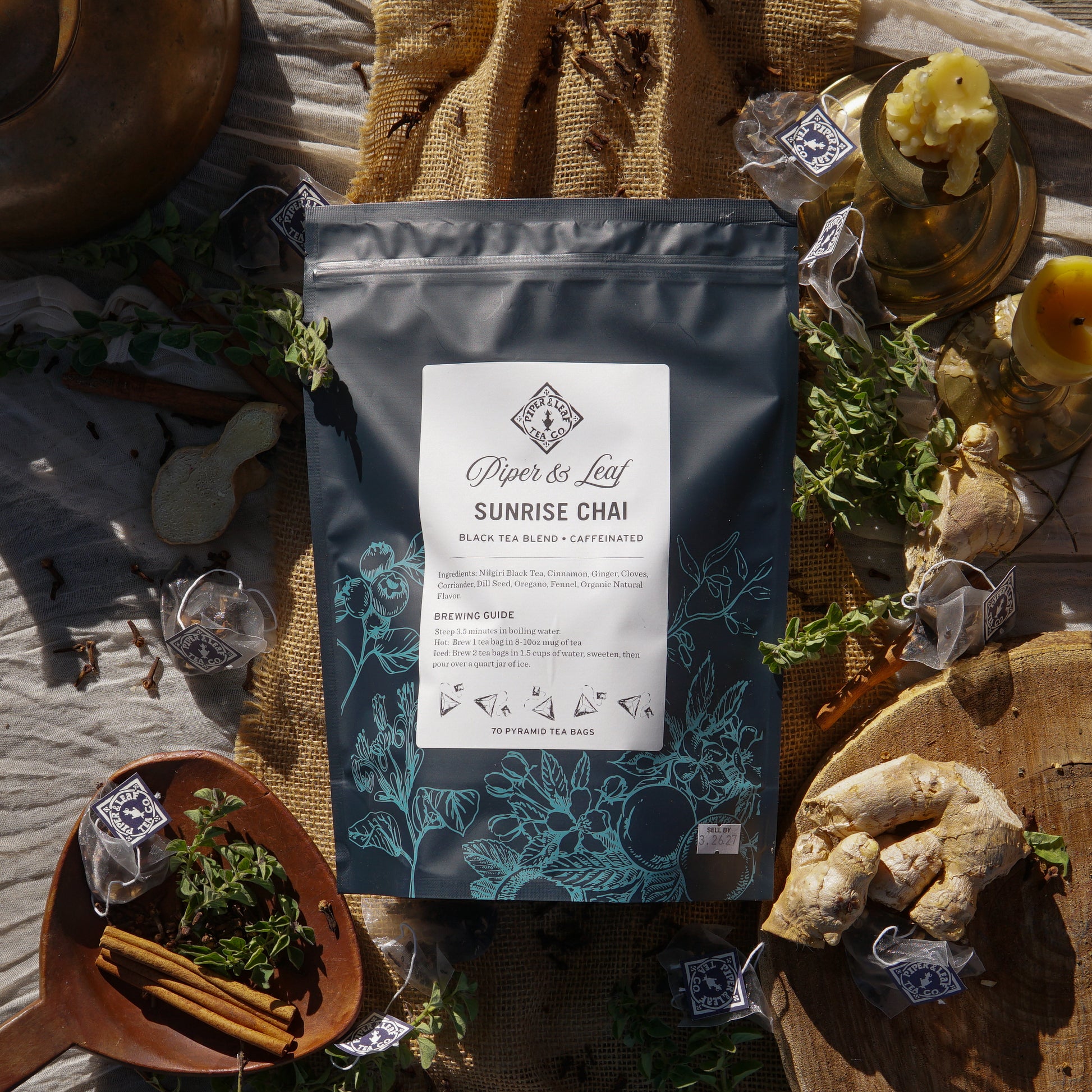 A bag of Piper & Leaf Tea Co. Sunrise Chai Bulk Sachets - 70ct Tea Bags is displayed on a rustic surface, surrounded by chai tea bags, fresh ginger, herbs, cinnamon sticks, and decorative items.
