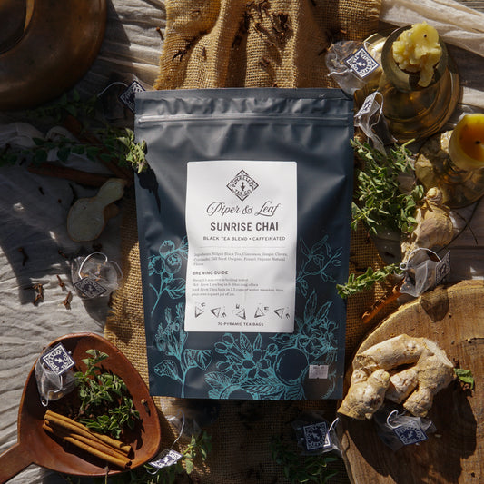 A bag of Piper & Leaf Tea Co. Sunrise Chai Bulk Sachets - 70ct Tea Bags is displayed on a rustic surface, surrounded by chai tea bags, fresh ginger, herbs, cinnamon sticks, and decorative items.