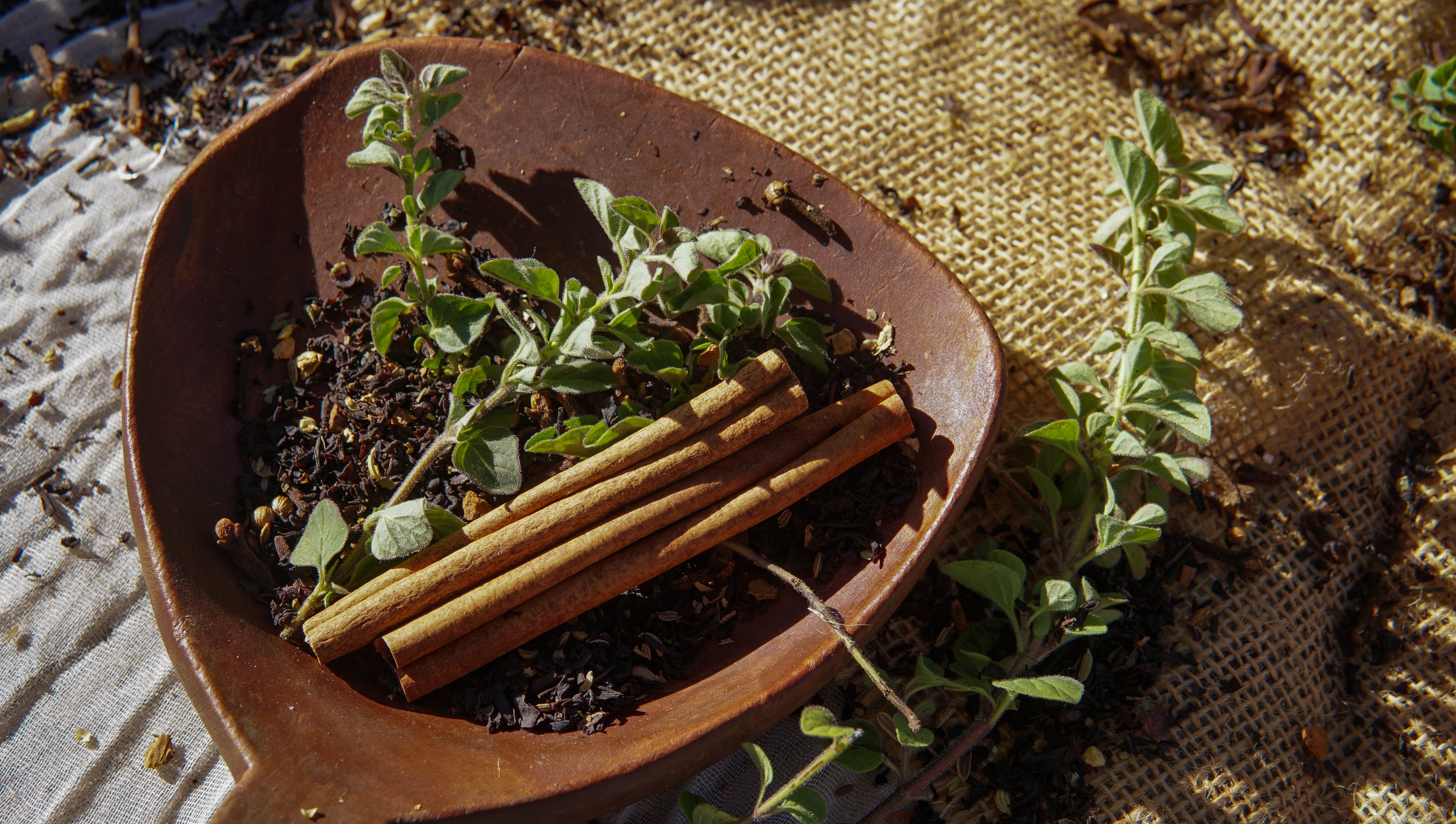 A brown dish holds three cinnamon sticks, a Piper & Leaf Tea Co. Sunrise Chai Muslin Bag of Loose Leaf Tea (15 servings), and fresh oregano sprigs, all arranged on burlap and cloth surfaces.