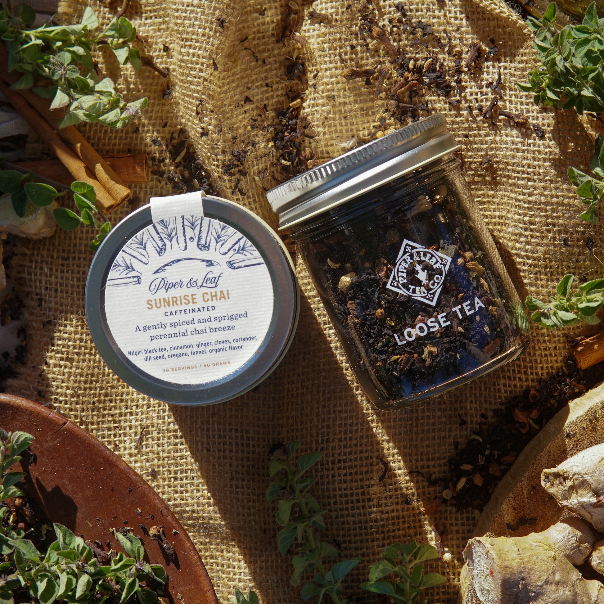 The Sunrise Chai Glass Jar of Loose Leaf Tea by Piper & Leaf Tea Co. sits on burlap with scattered tea leaves, cinnamon sticks, and ginger root, highlighting this vibrant spiced black tea blend. Contains 30 servings.